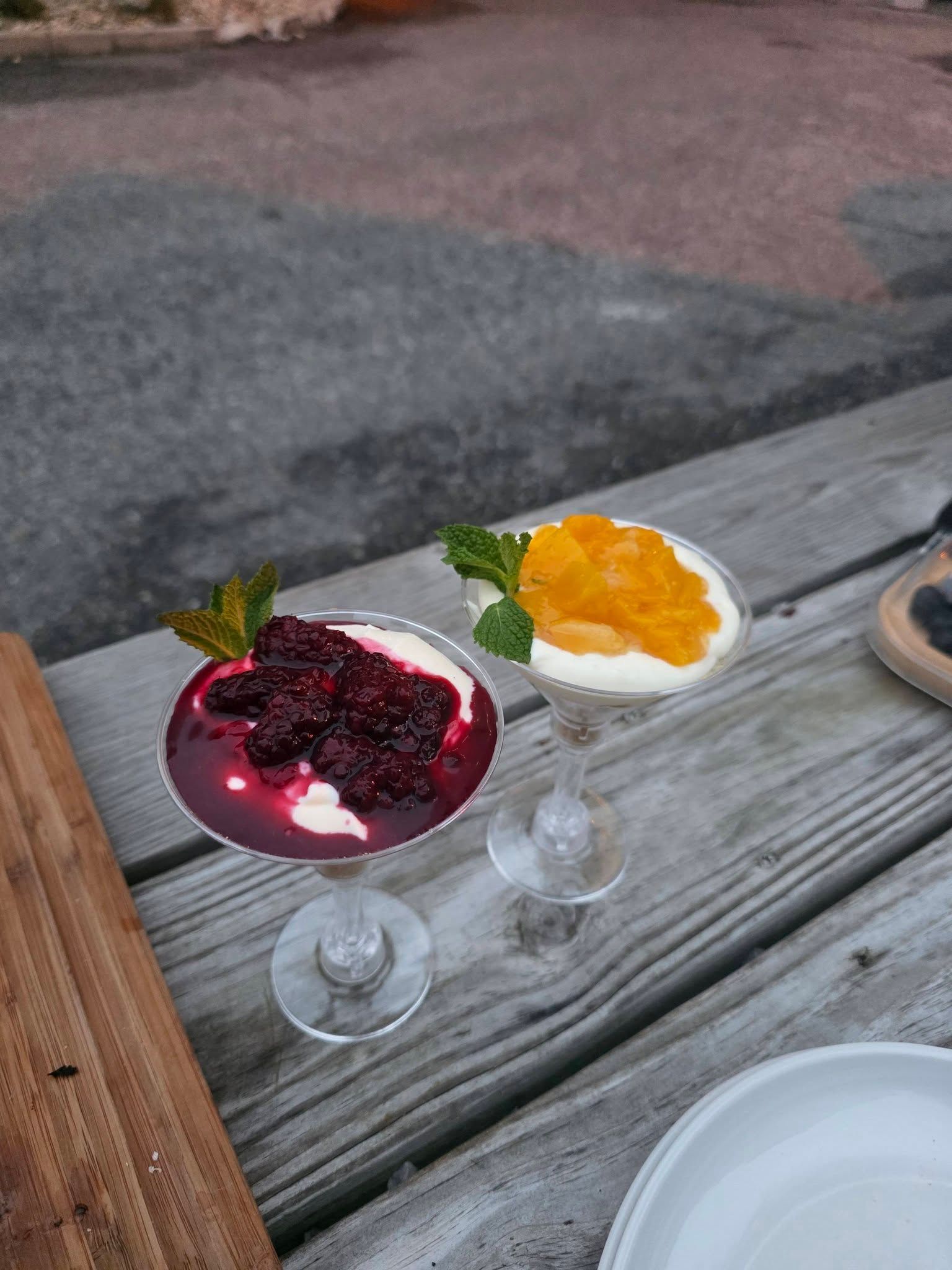 Two dessert glasses on a weathered picnic table, one with purple berry topping and one with orange topping.