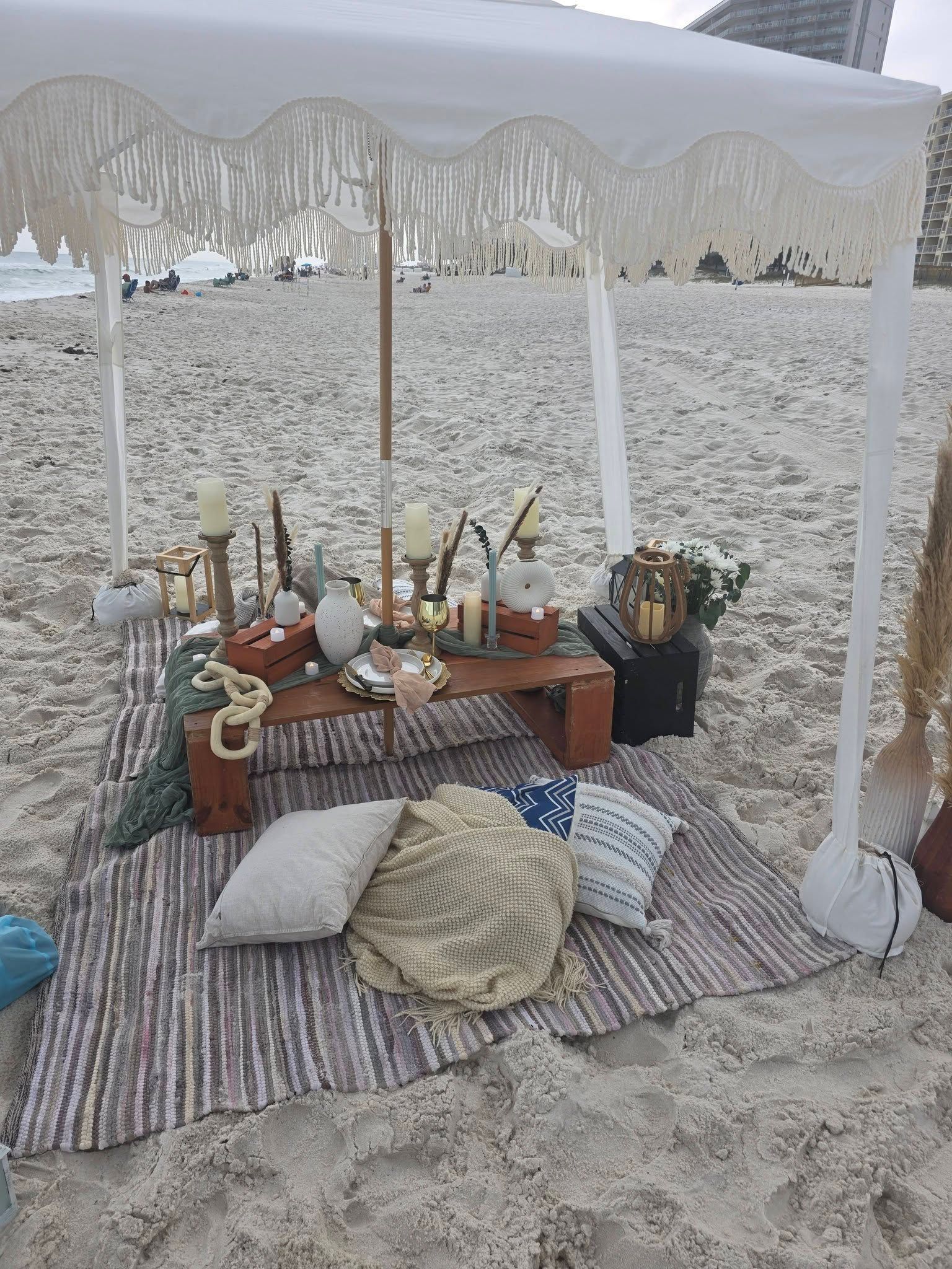 Beach picnic setup under a white canopy with pillows, rugs, and food on the sand