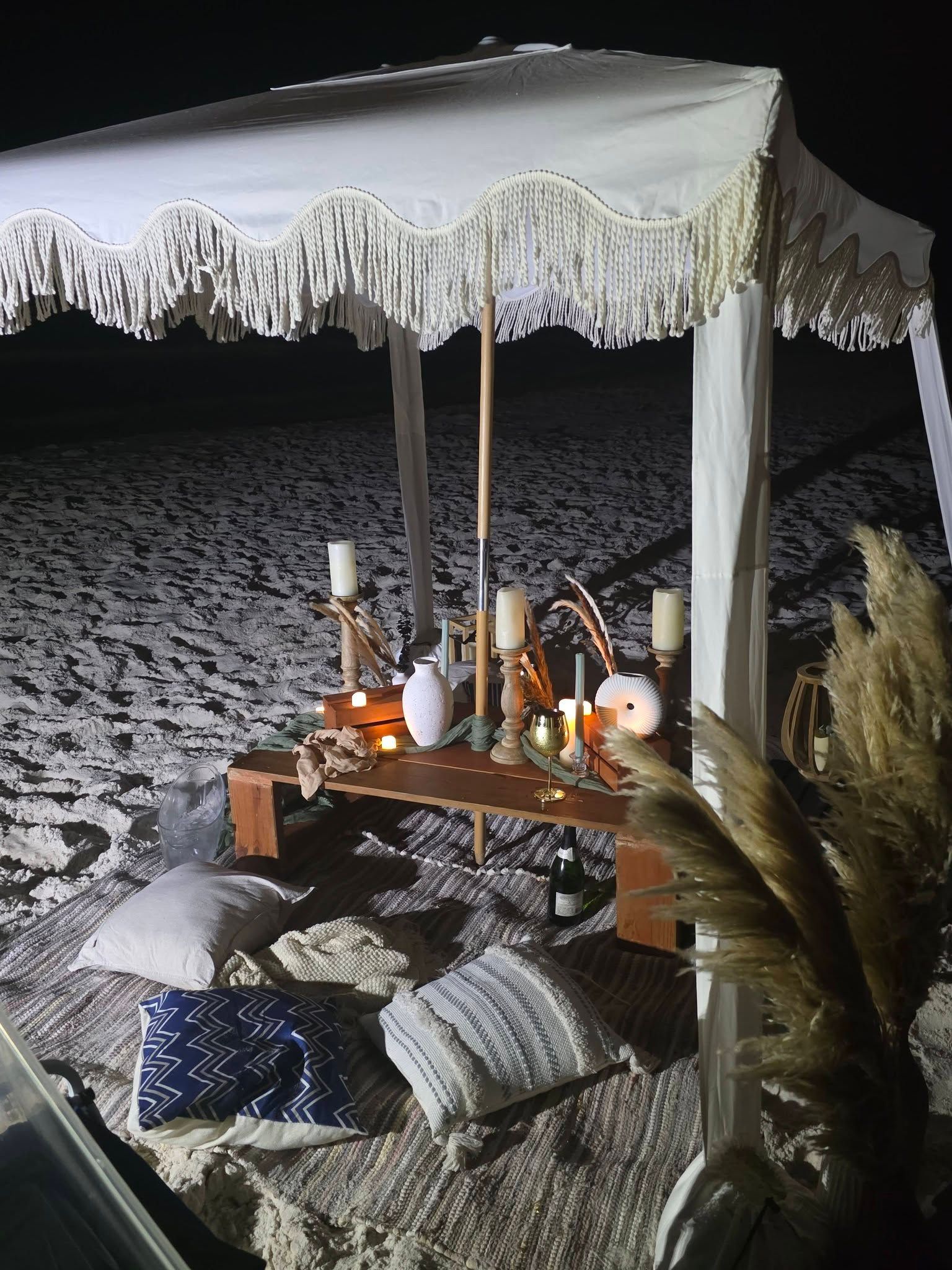 Beachside candlelit setup under a white canopy at night, with cushions and low table on the sand.