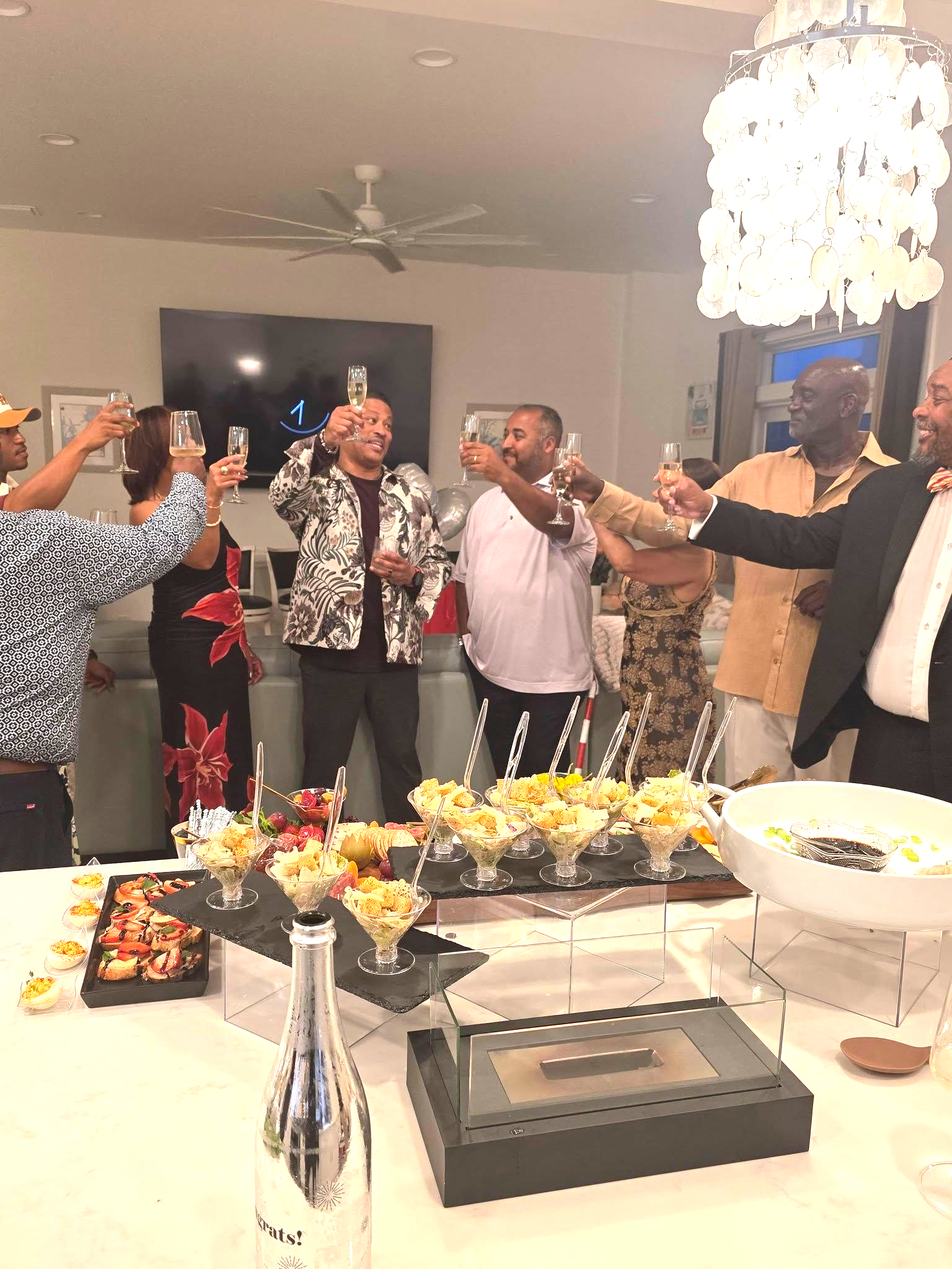 Guests toast at a buffet table in a decorated banquet hall with chandelier lighting.