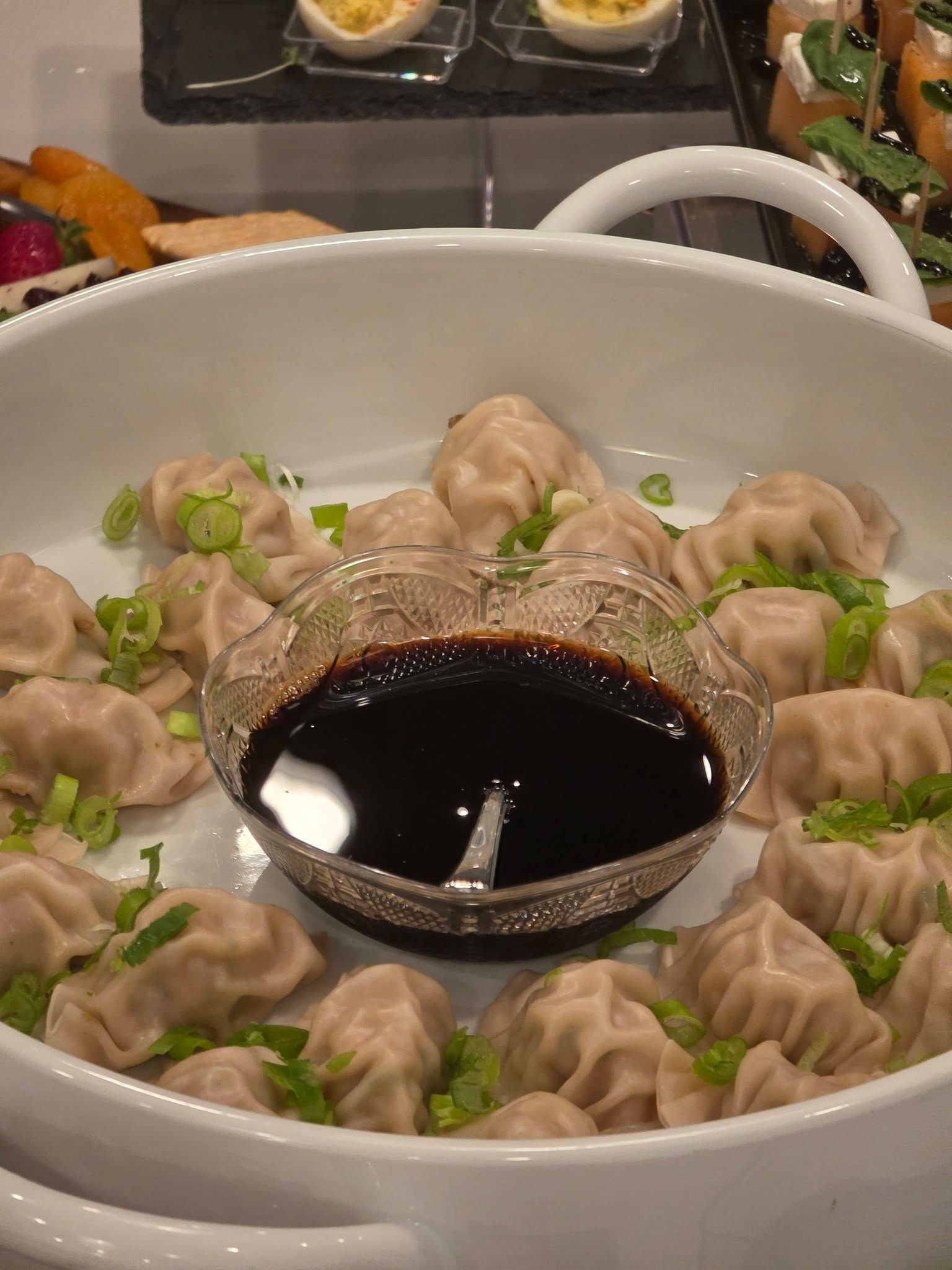 Dumplings arranged around a bowl of dark dipping sauce on a white serving dish with chopped scallions.