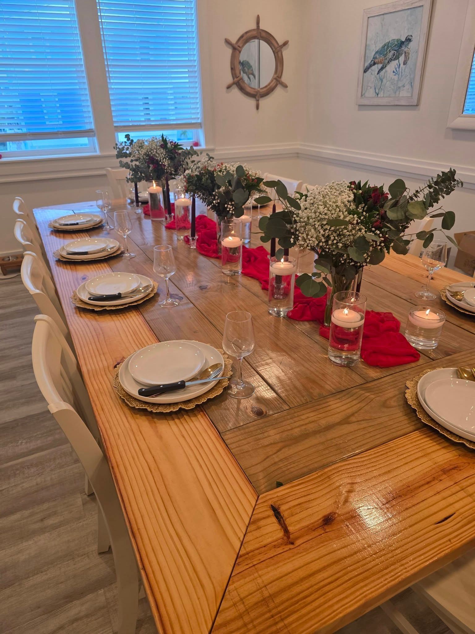 Decorated dining table with red candles, greenery, plates, and white chairs in a bright room