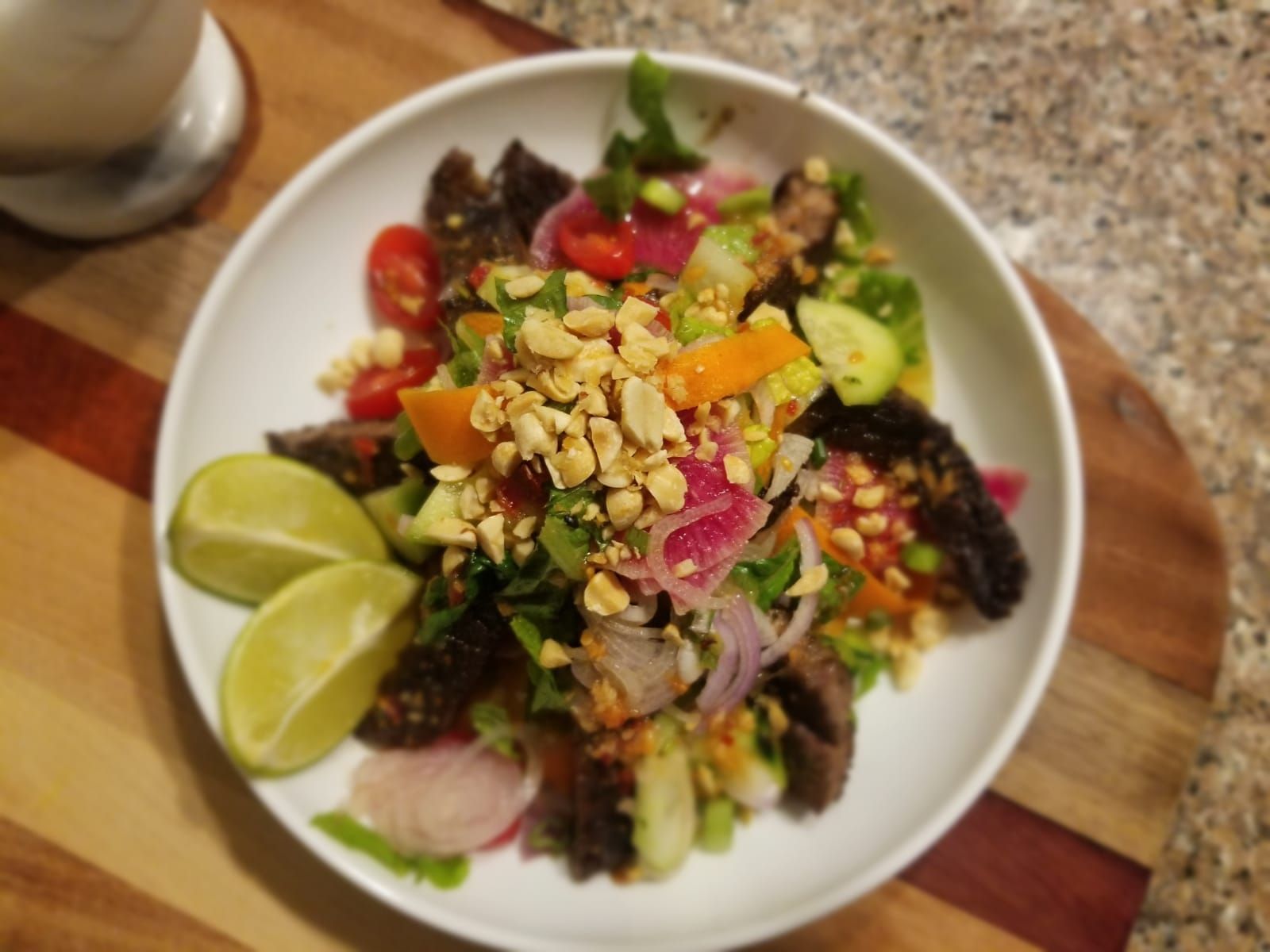 Bowl of colorful salad with lime wedges on a wooden board, topped with chopped nuts and vegetables