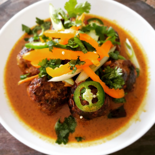 Bowl of spicy meatballs in red curry, topped with herbs, carrot strips, and sliced green chili.