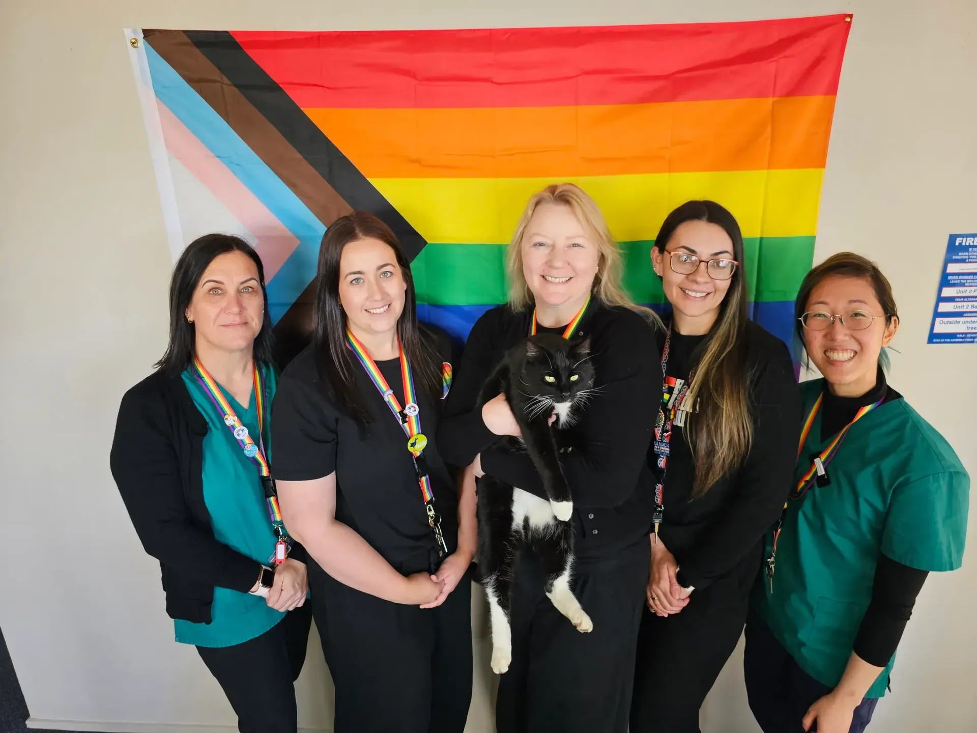 Five people wearing rainbow lanyards stand together in front of a progress pride flag, one of them holding a black and white cat