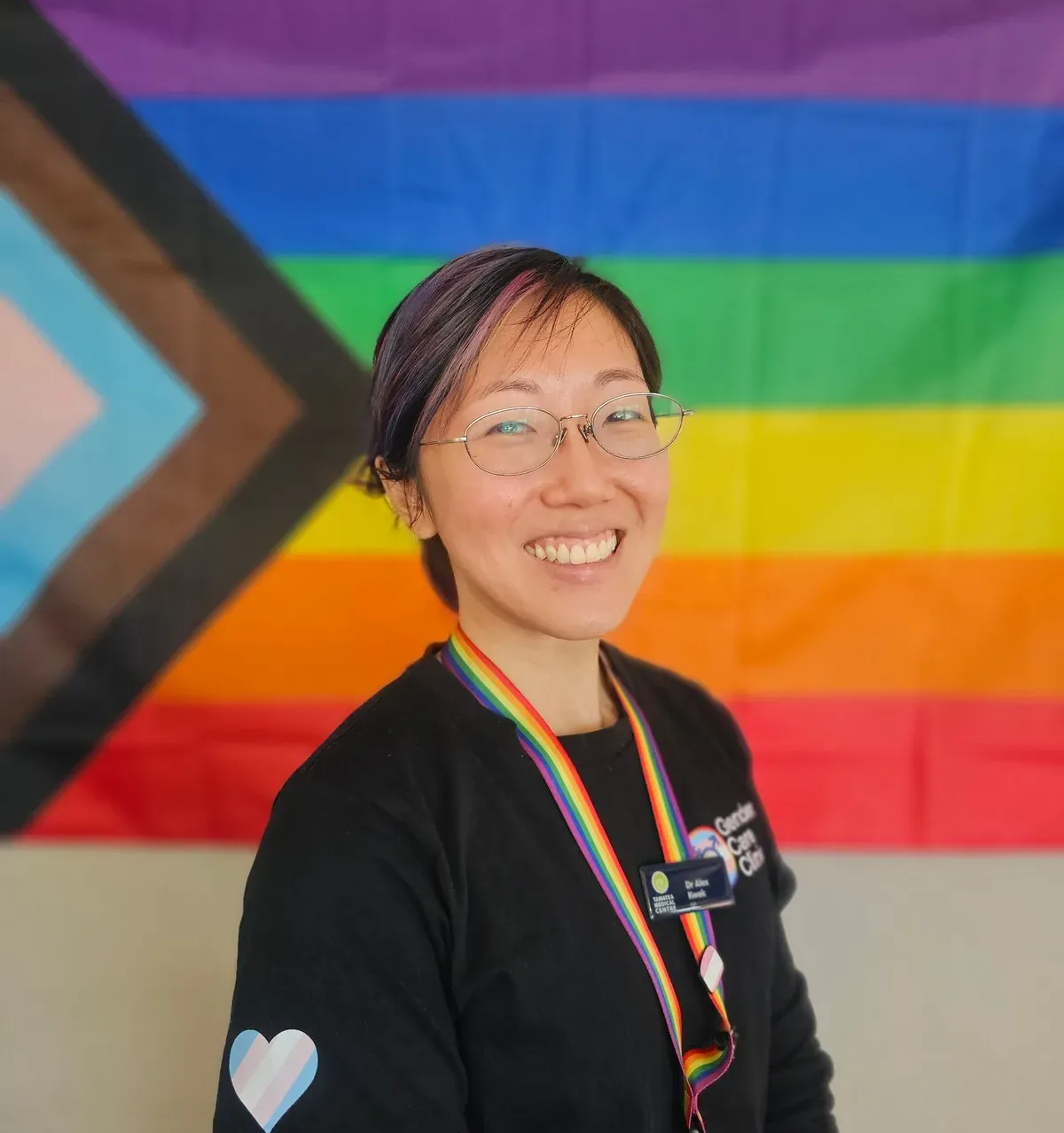 Dr Alex Kwek smiling, with short dark hair, glasses and a rainbow lanyard, wearing a black Gender Care Clinic T-shirt in front of a progress pride flag