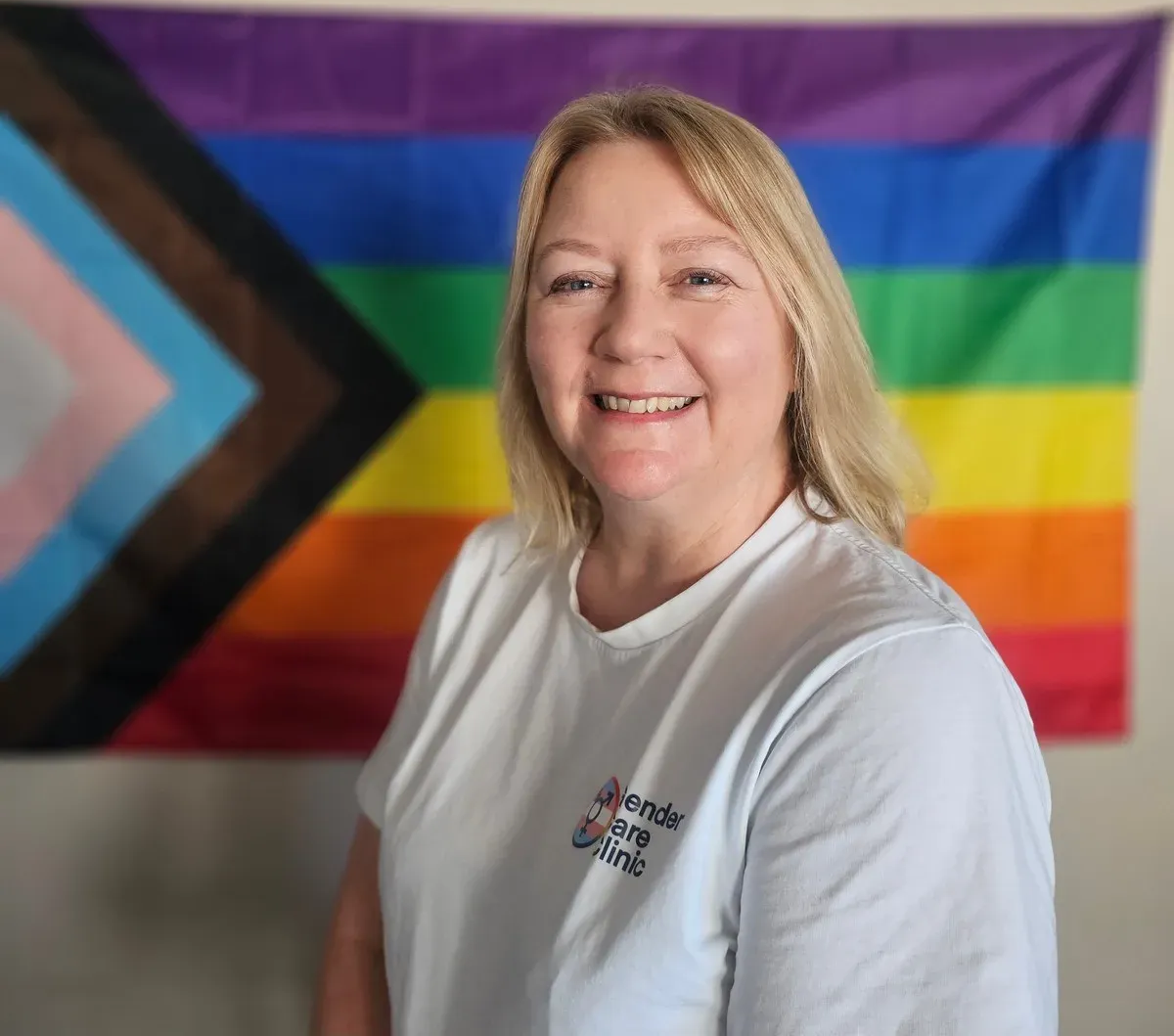 Dr Elizabeth McElrea smiling, wearing a white Gender Care Clinic T-shirt in front of a progress pride flag