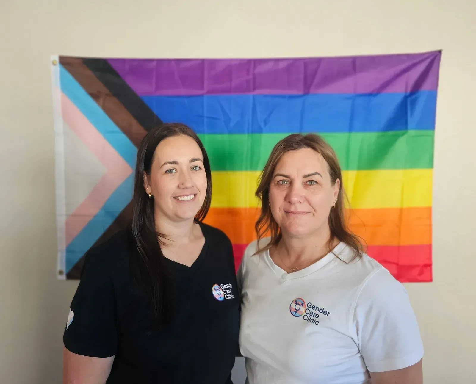 Nurses Natalie and Jenna standing side by side in front of a progress pride flag, one in a black and one in a white Gender Care Clinic T-shirt