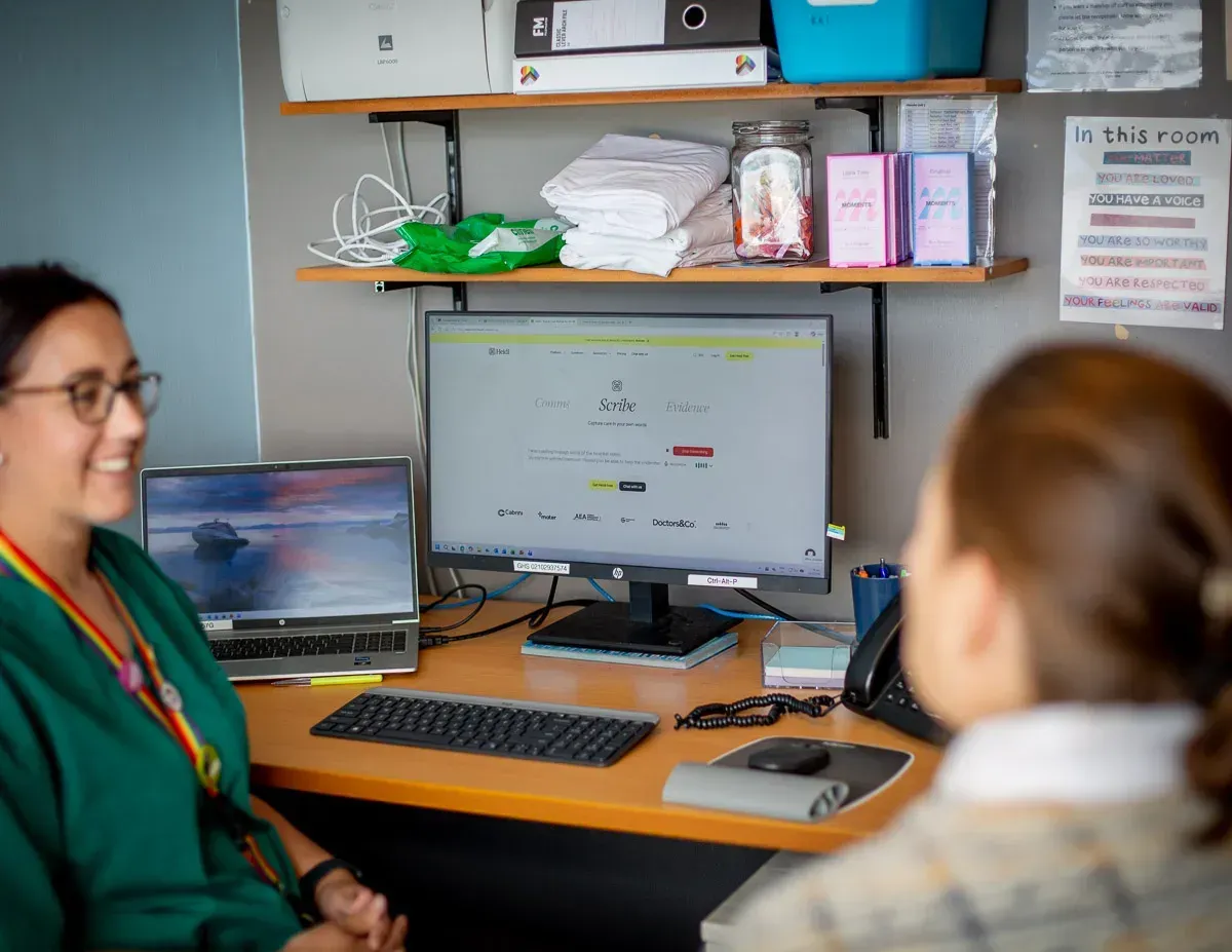 A consultation room where a smiling person in a green top and rainbow lanyard talks with someone facing away from the camera, across a desk with a computer