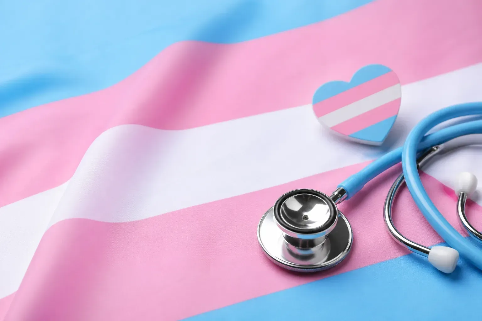 A stethoscope and a small heart in transgender pride flag colours resting on a transgender pride flag
