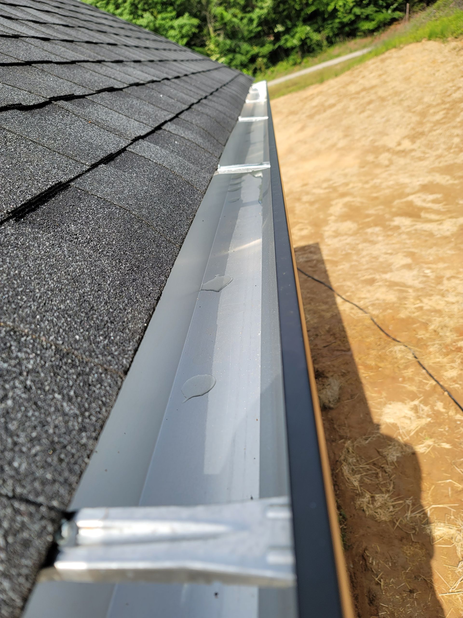 Gray asphalt shingle roof with silver gutter, against a brown, dirt-like surface, outdoors.