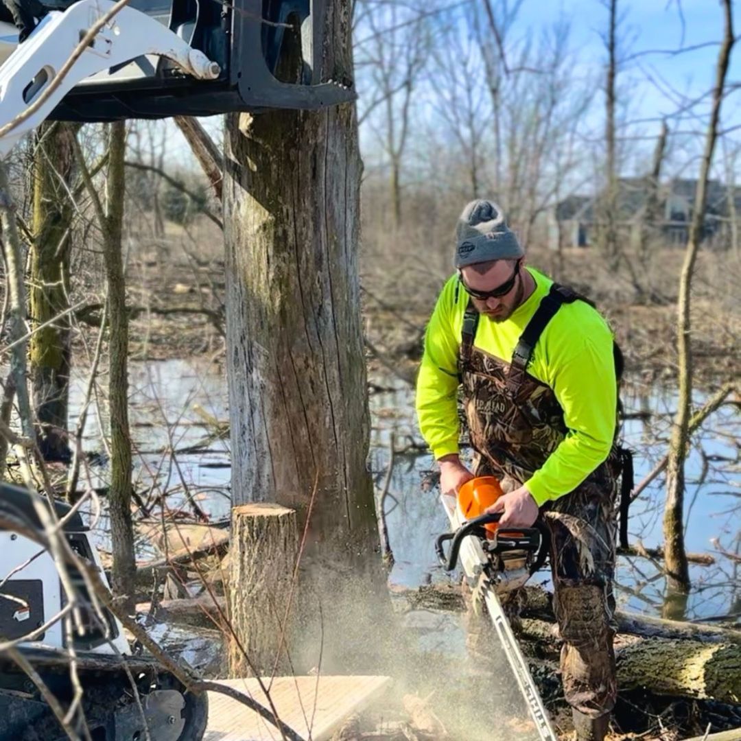 Man cutting tree with chainsaw near flooded area, wearing safety gear.