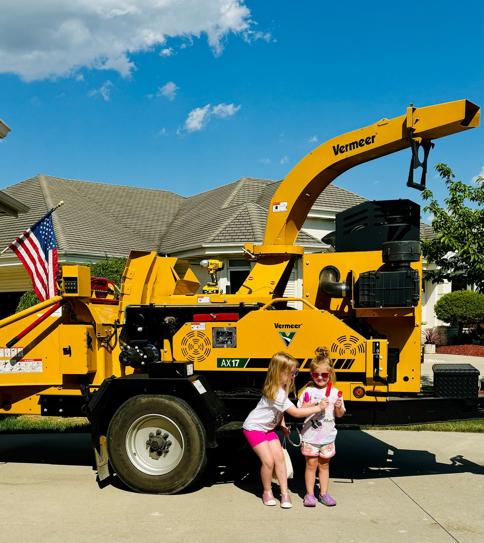 Two children stand near a large yellow wood chipper, American flag attached, blue sky.