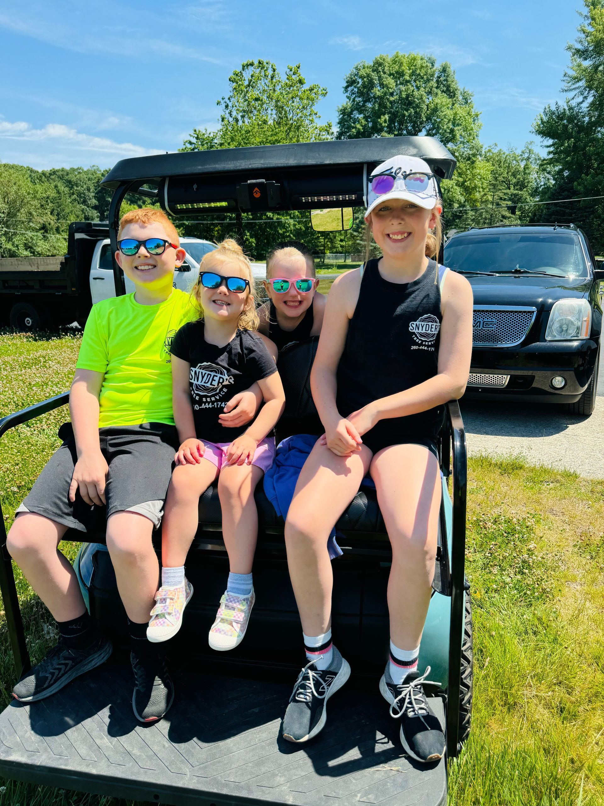 Four children wearing sunglasses sit on a golf cart on a sunny day.