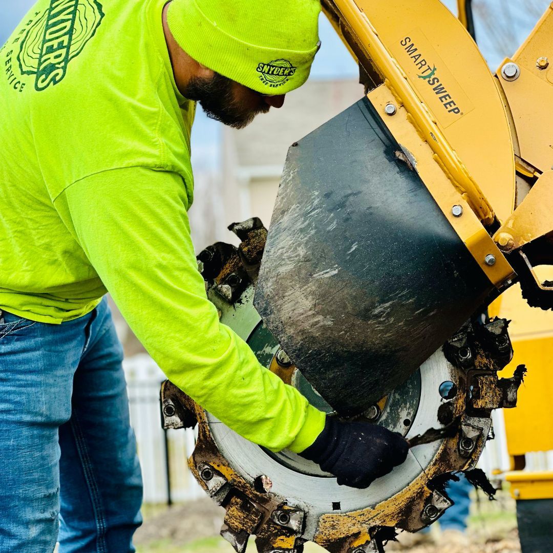 Man in neon green shirt works on stump grinder; outdoors, sunny.