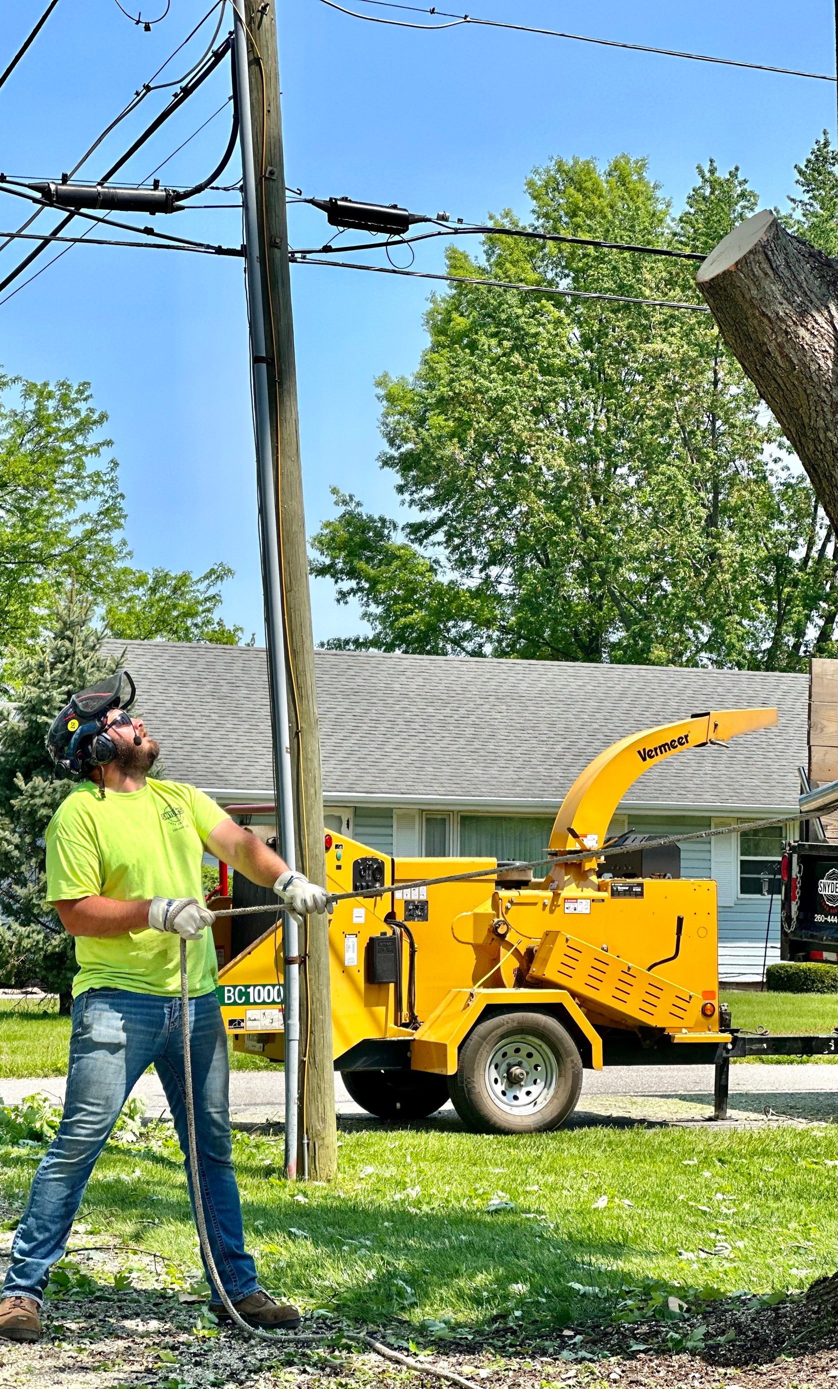 Man in safety gear pulls rope near power lines and wood chipper.