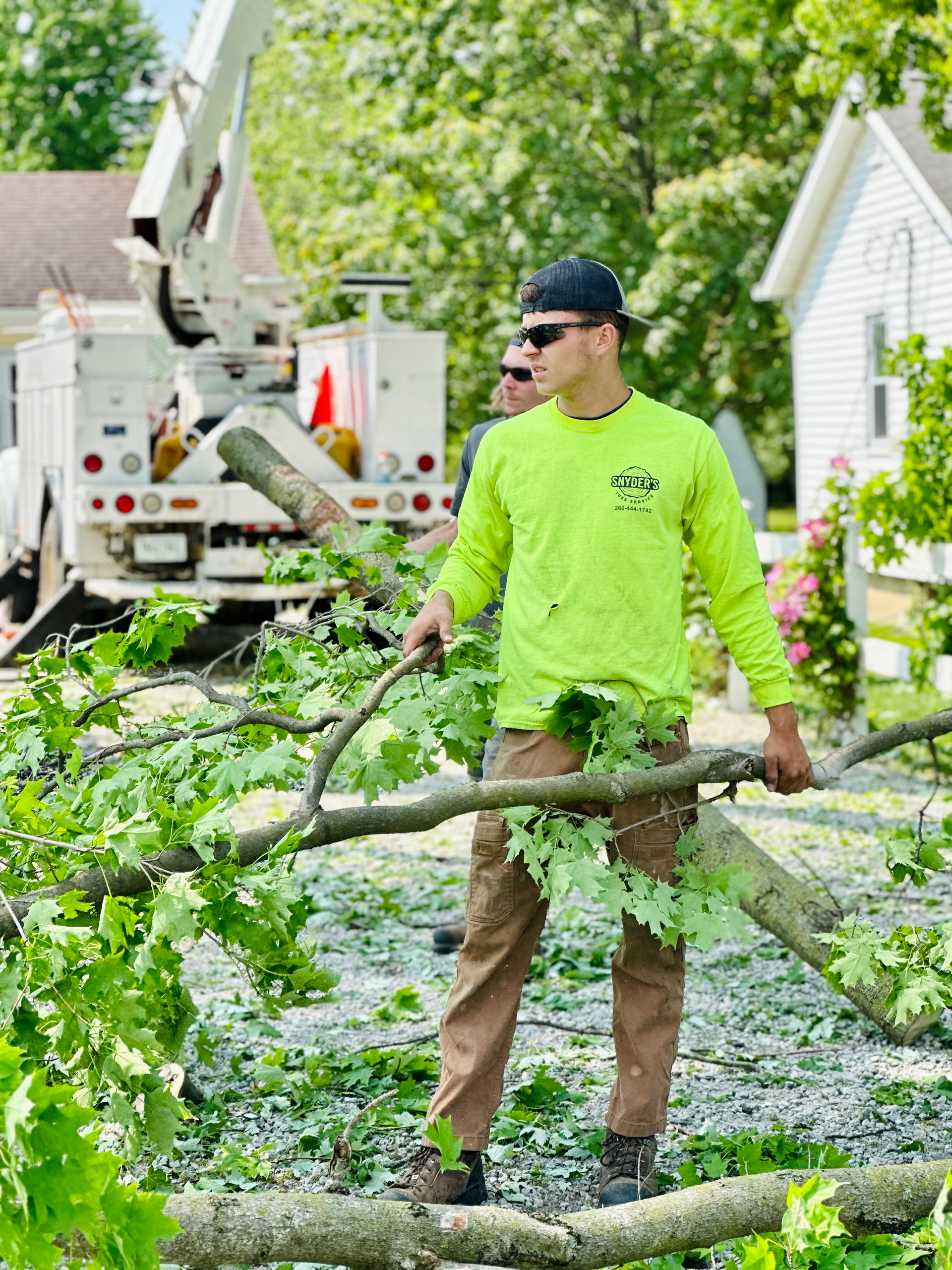 Arborist holding tree branch, standing in front of a truck, wearing neon green shirt and khaki pants.