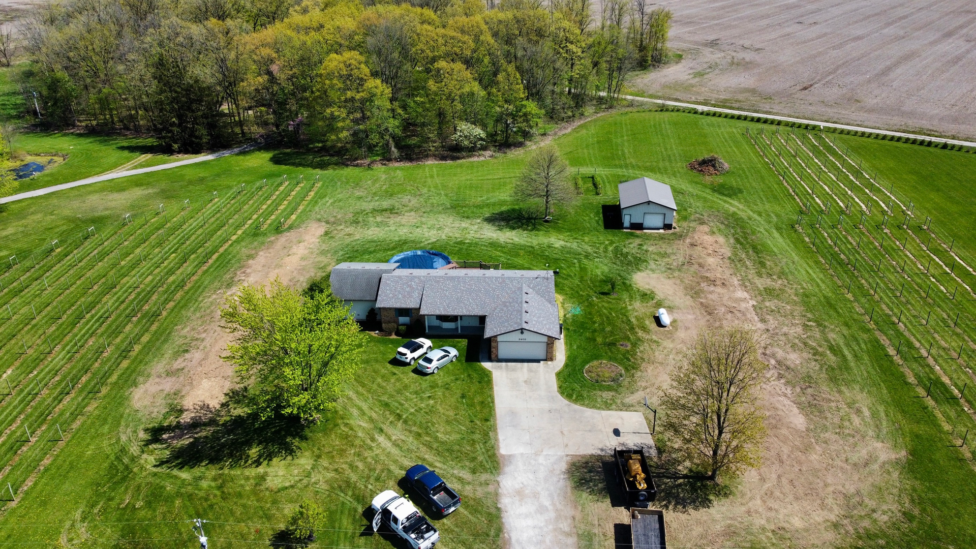 Aerial view of a house with a driveway, garage, and adjacent outbuildings on a grassy hill; surrounding farmland.