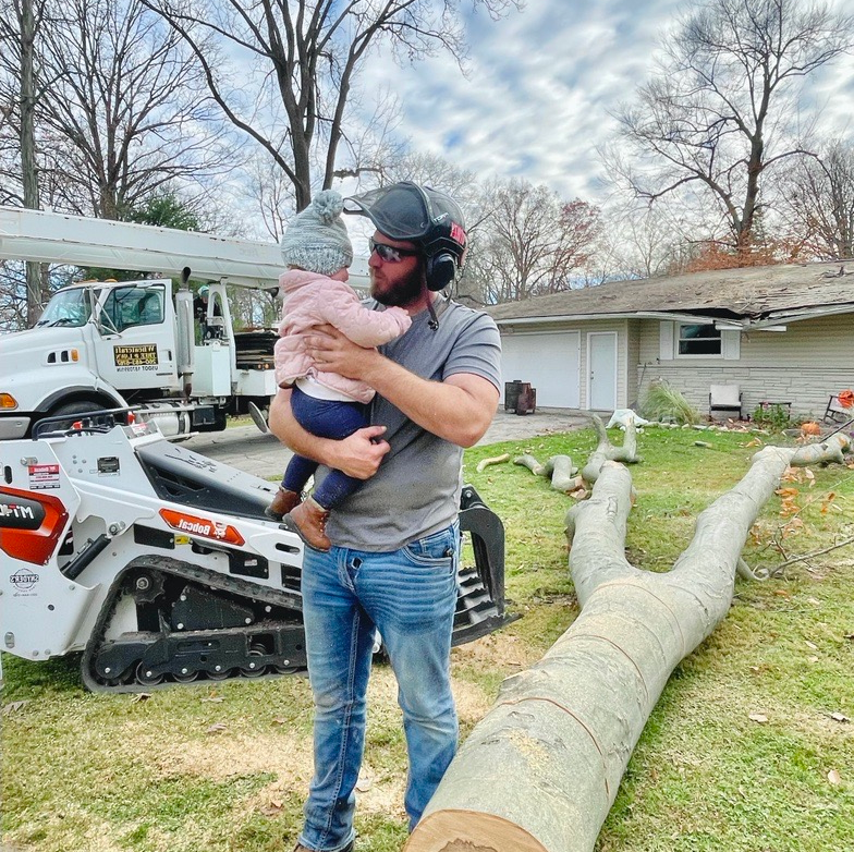 Man holding a baby, wearing work gear, next to tree equipment and cut log.