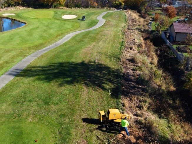 A worker operating a yellow machine on a grassy hillside next to a golf course path.