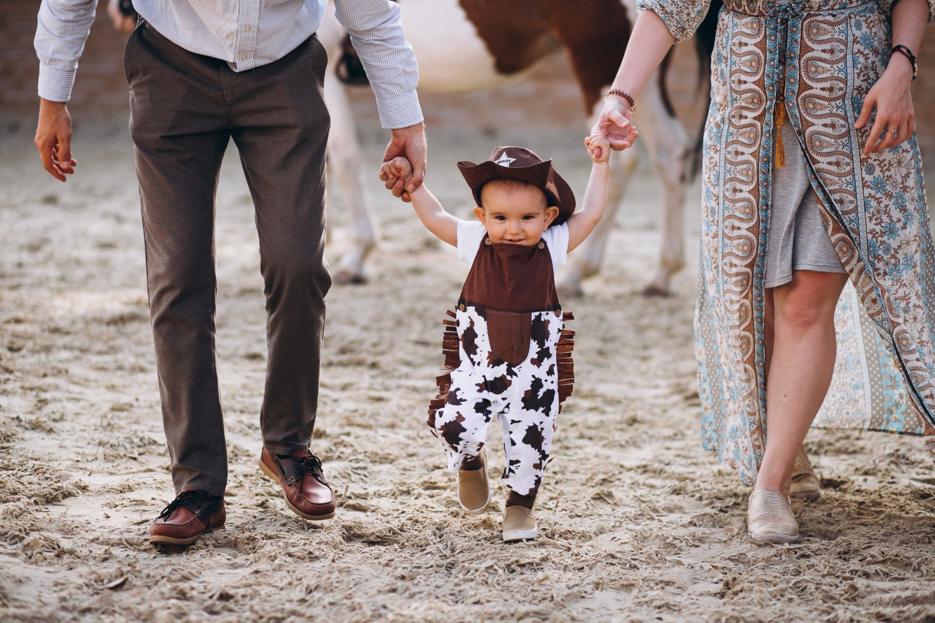 Family walking on a beach, holding a toddler’s hands as they take steps in the sand.