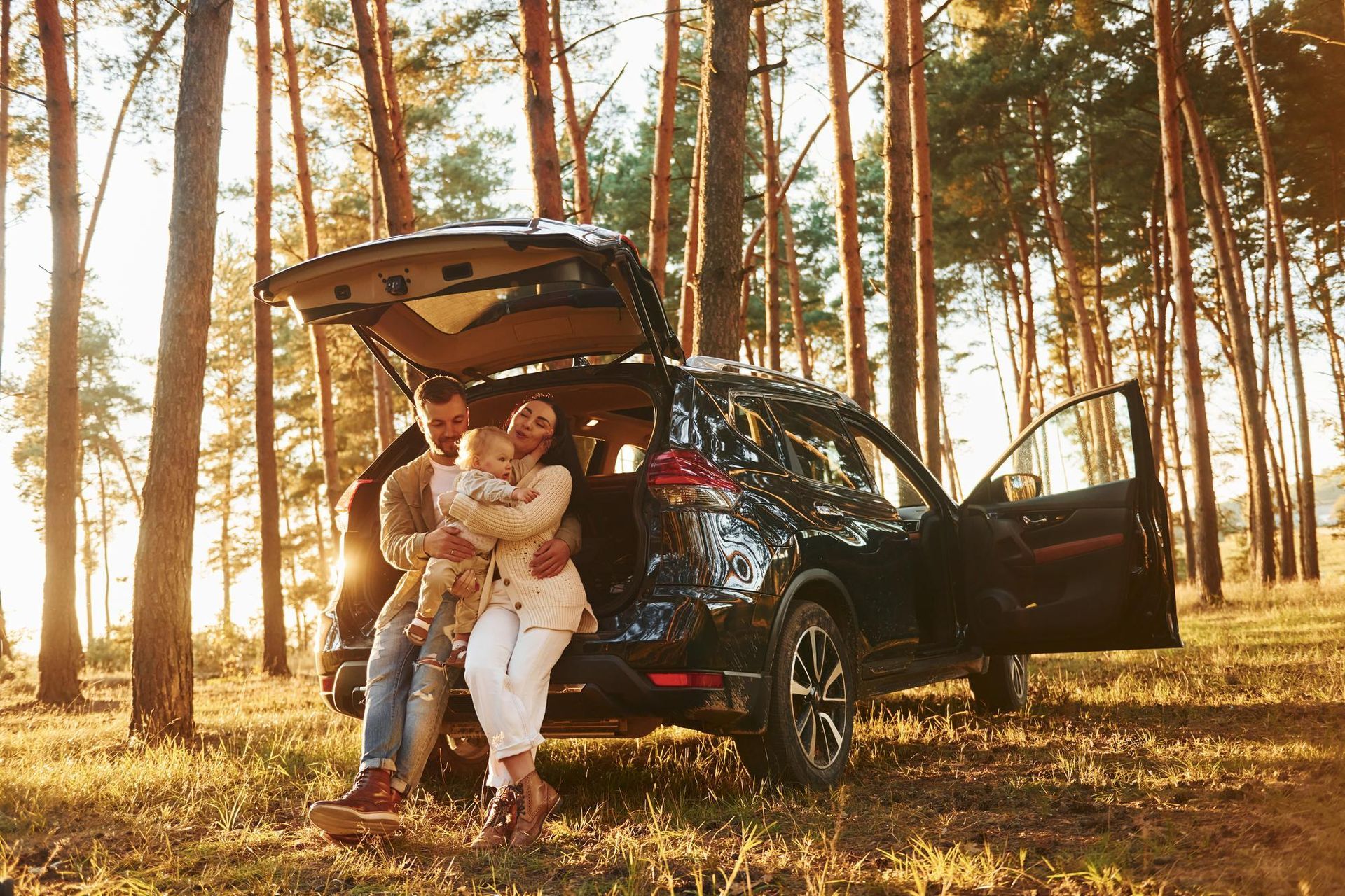 Couple with child beside open SUV in a sunlit forest clearing, enjoying a picnic outdoors