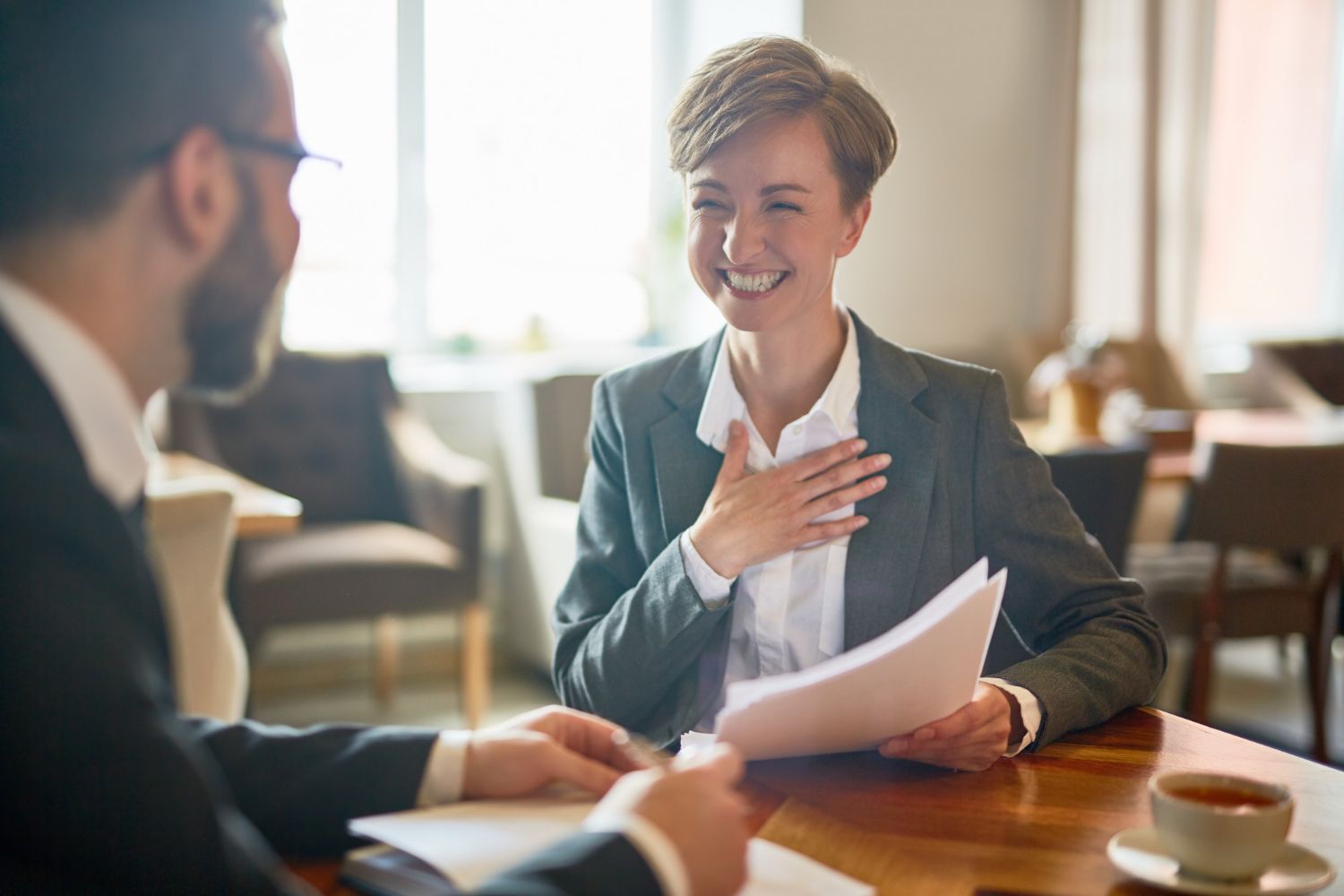 Two coworkers smiling and talking at a café table, one holding papers and the other gesturing