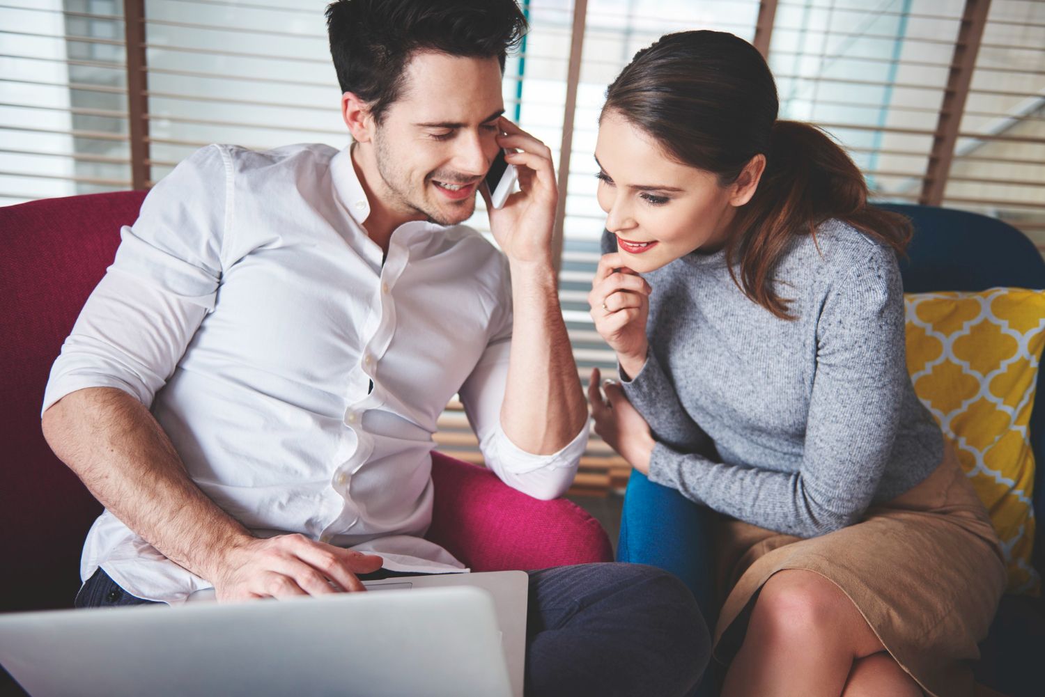 Two people smiling and leaning over a laptop on a couch, discussing something together in a cozy room