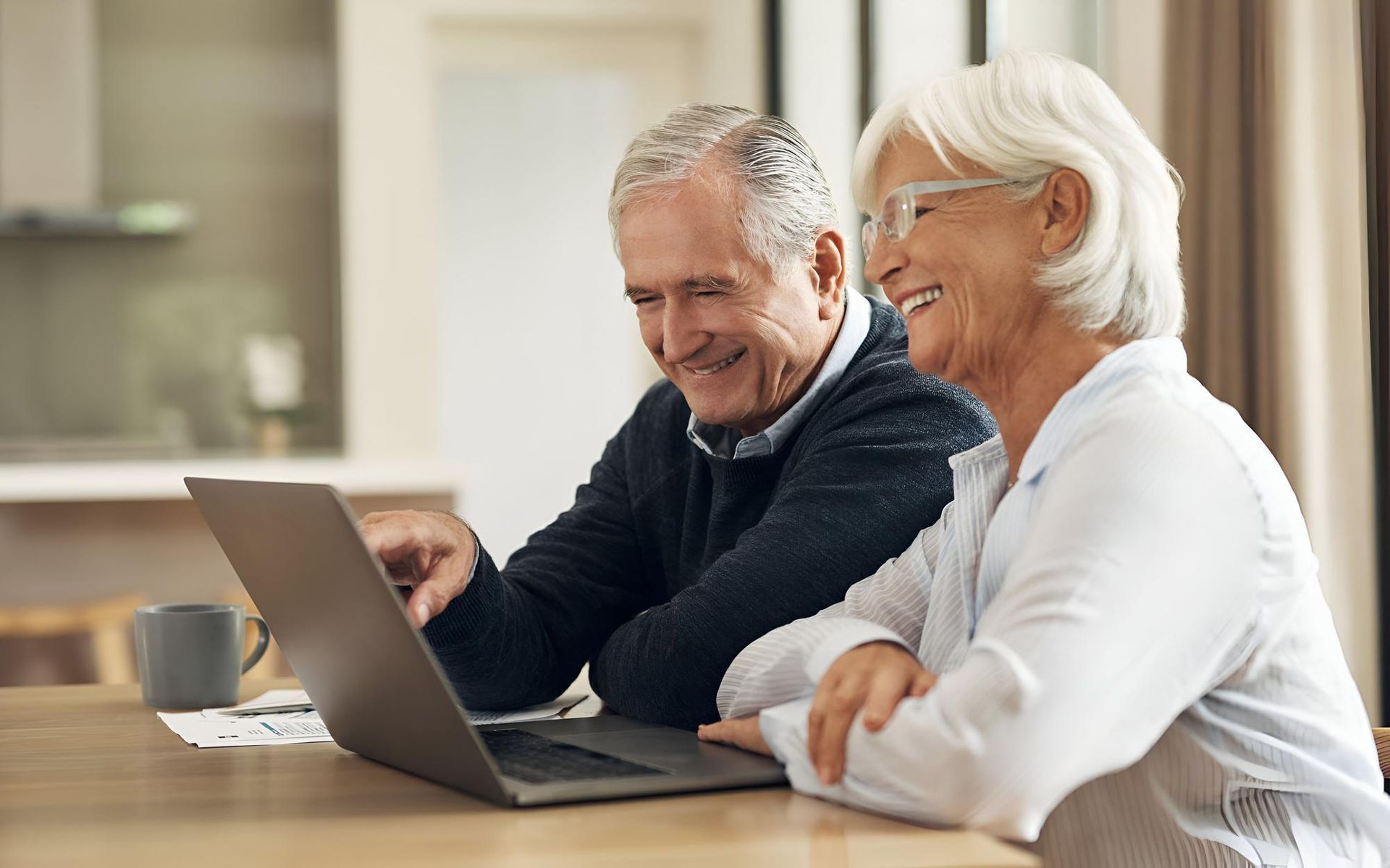 Two smiling people using a laptop together at a table in a bright kitchen