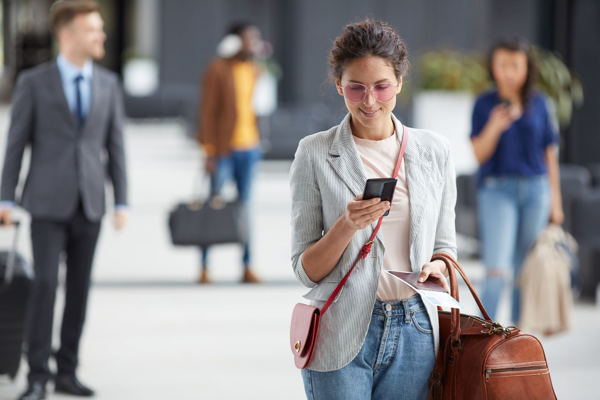 Woman with glasses texting while walking through a busy airport terminal, carrying a brown bag and striped tote