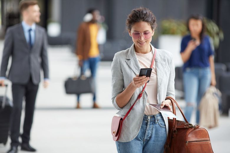 Woman with glasses texting while walking through a busy airport terminal, carrying a brown bag and striped tote