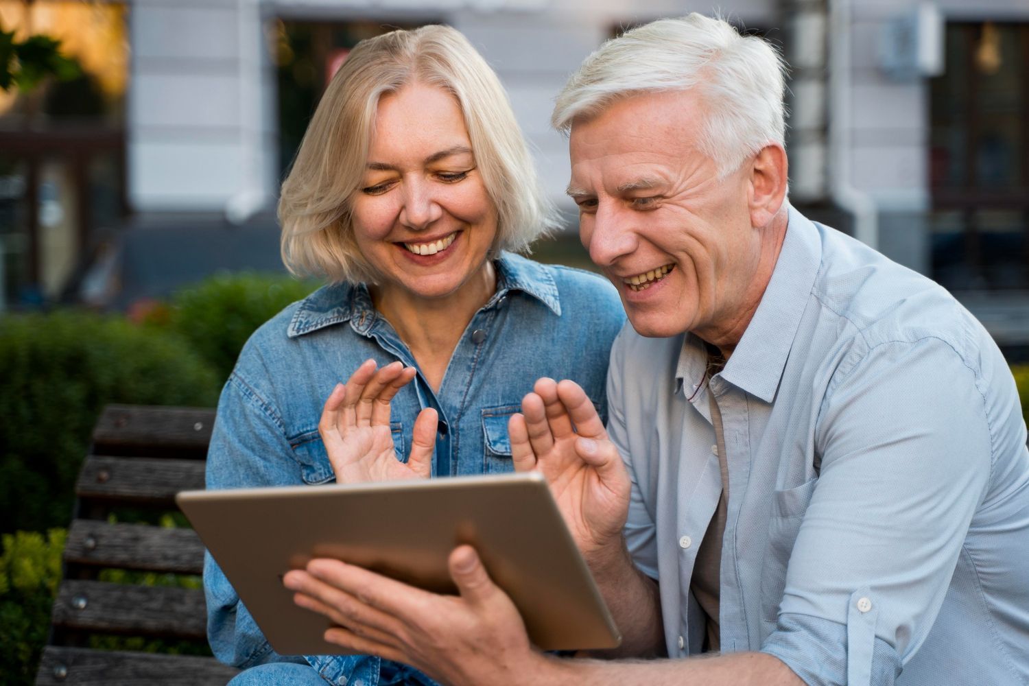 Two smiling people using a tablet on a park bench outdoors
