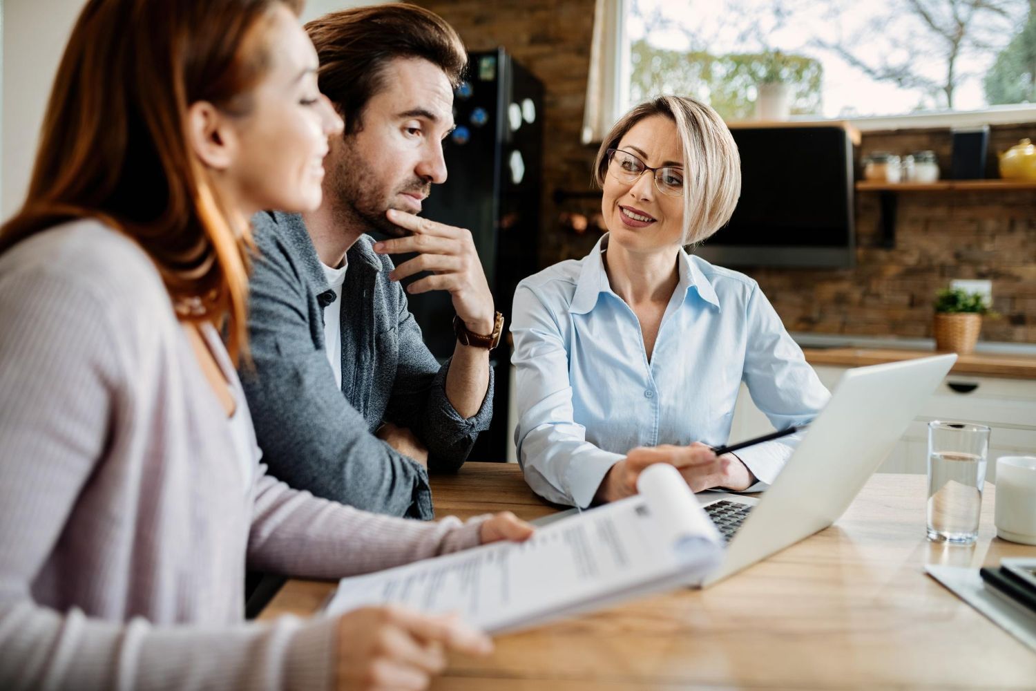 Three coworkers in a meeting around a laptop at a wooden table, discussing documents in a bright office.