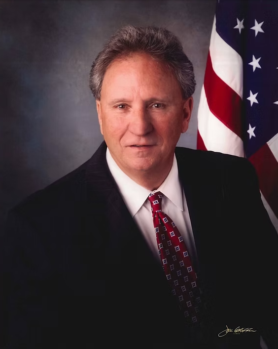 Formal portrait of a man in a suit and red tie, with an American flag in the background.