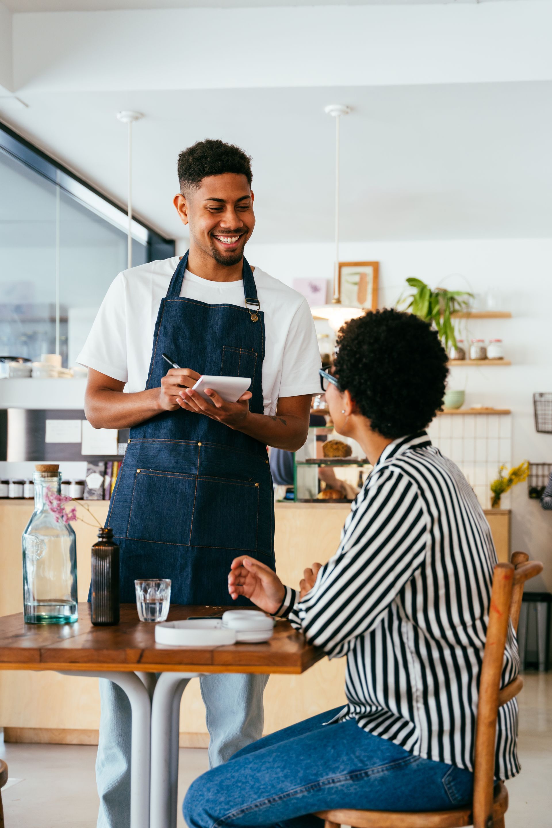 Barista serving a seated customer in a bright café, smiling and holding a receipt