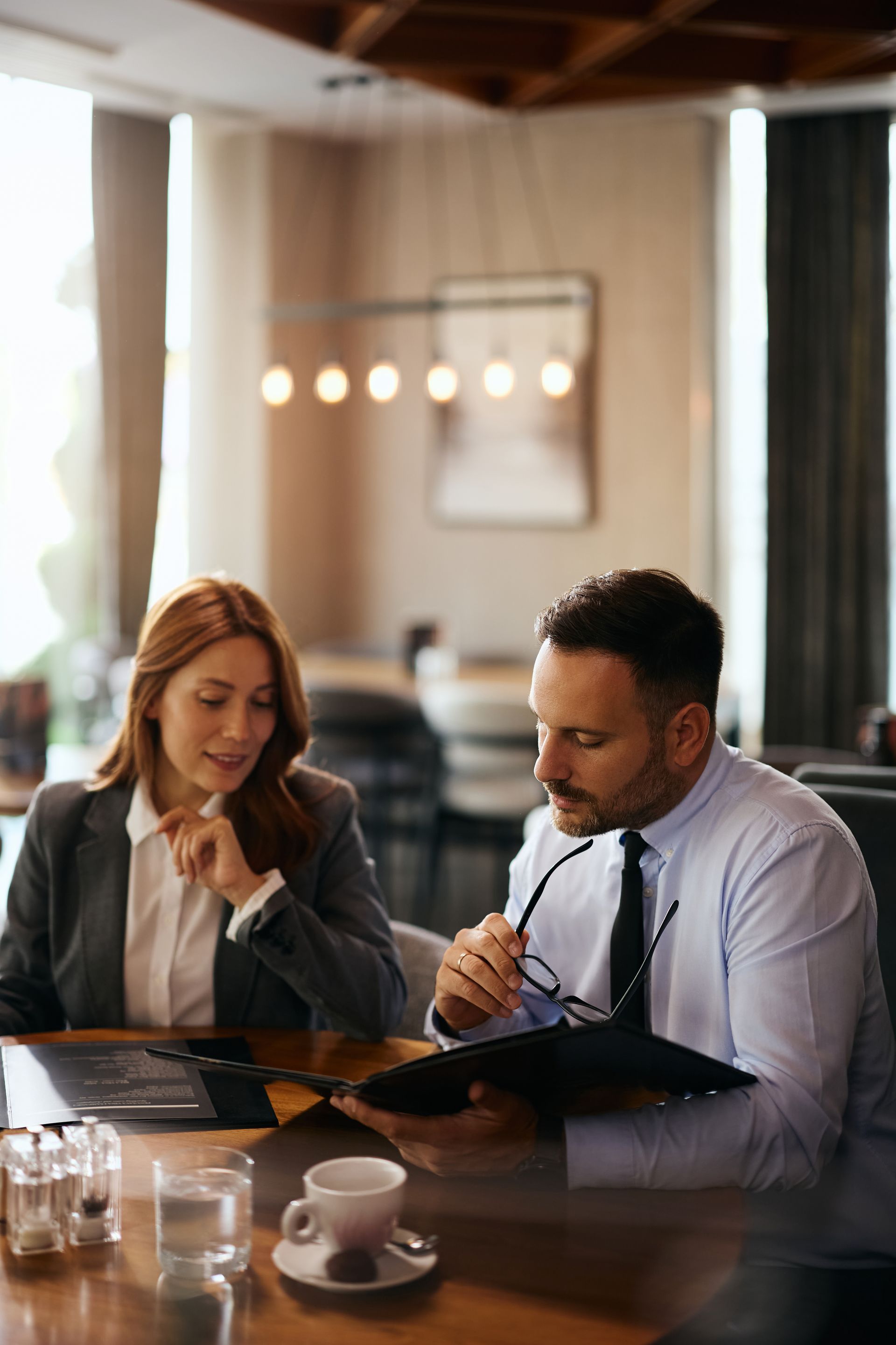 Two people reviewing a menu at a restaurant table, with coffee cups and warm pendant lights in the background.