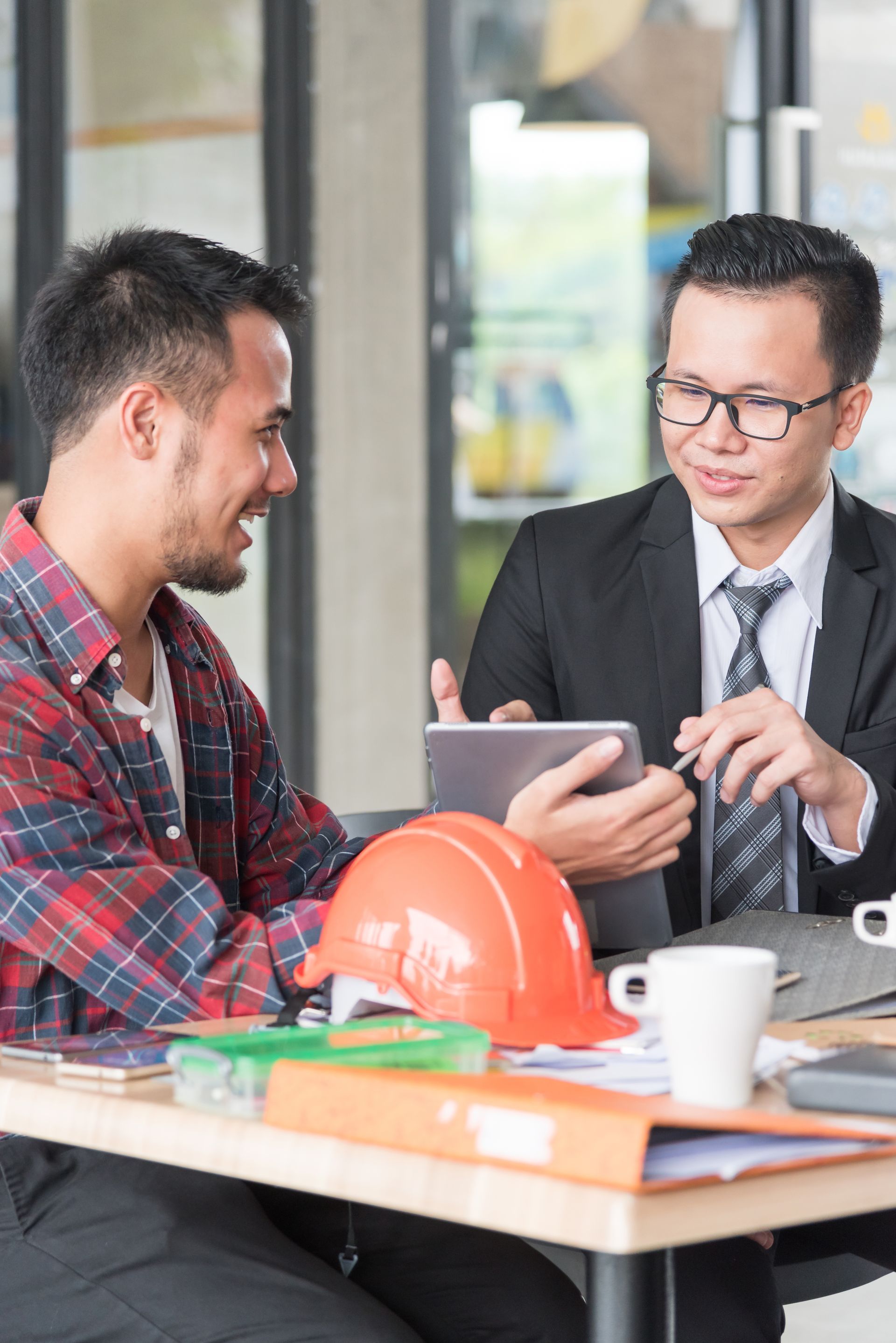 Two men discussing a tablet at a desk with a hard hat and coffee cup in a bright office