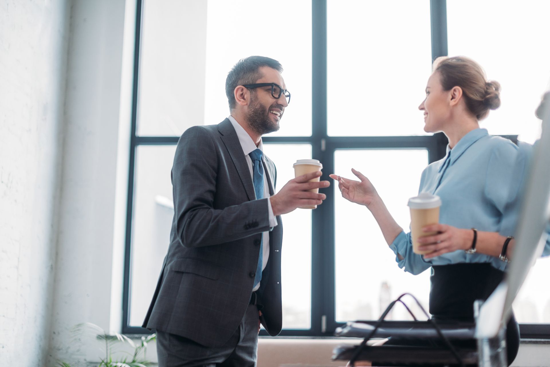Two coworkers talking in a bright office, holding coffee cups by a large window.