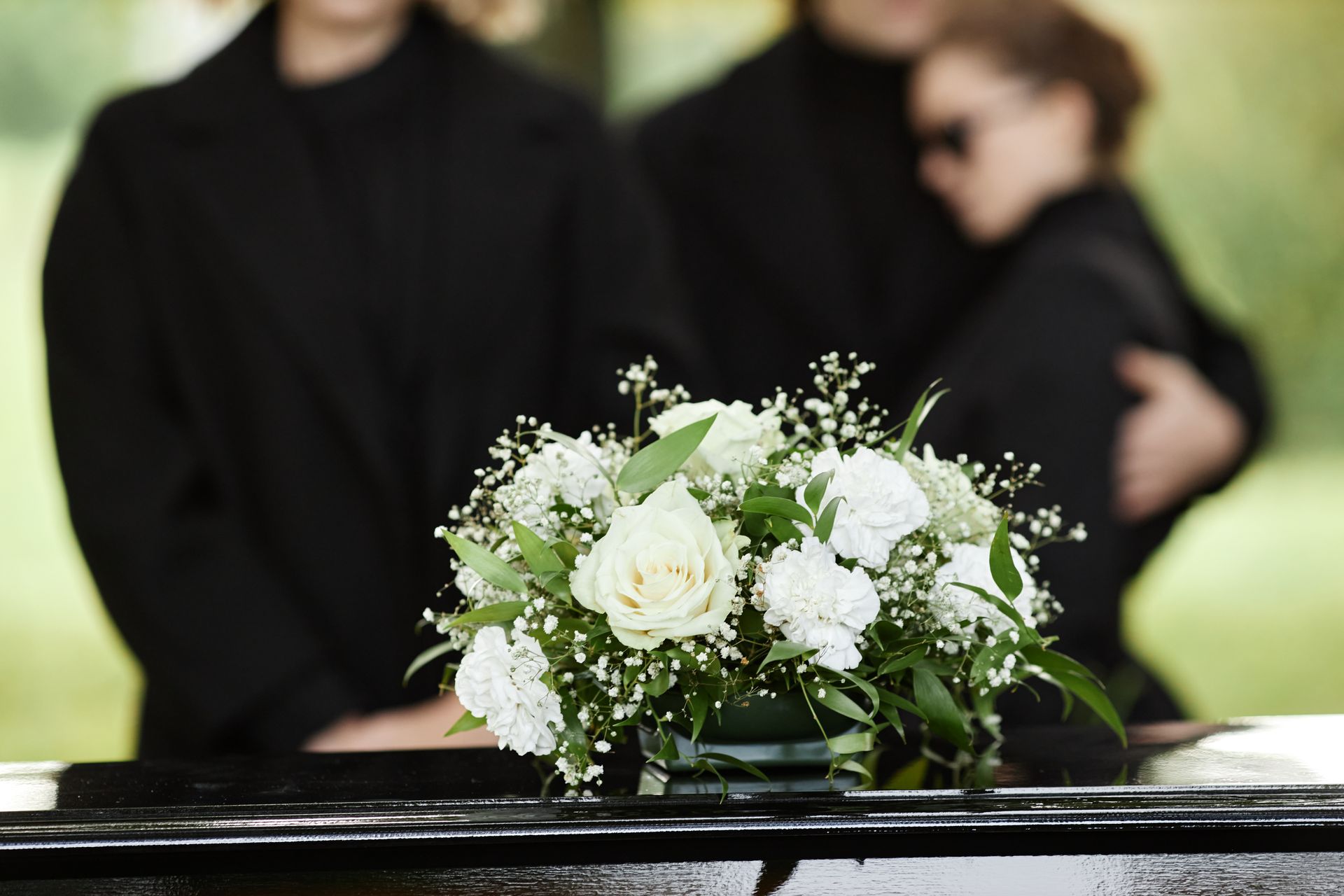 White floral arrangement on a casket with mourners blurred in the background