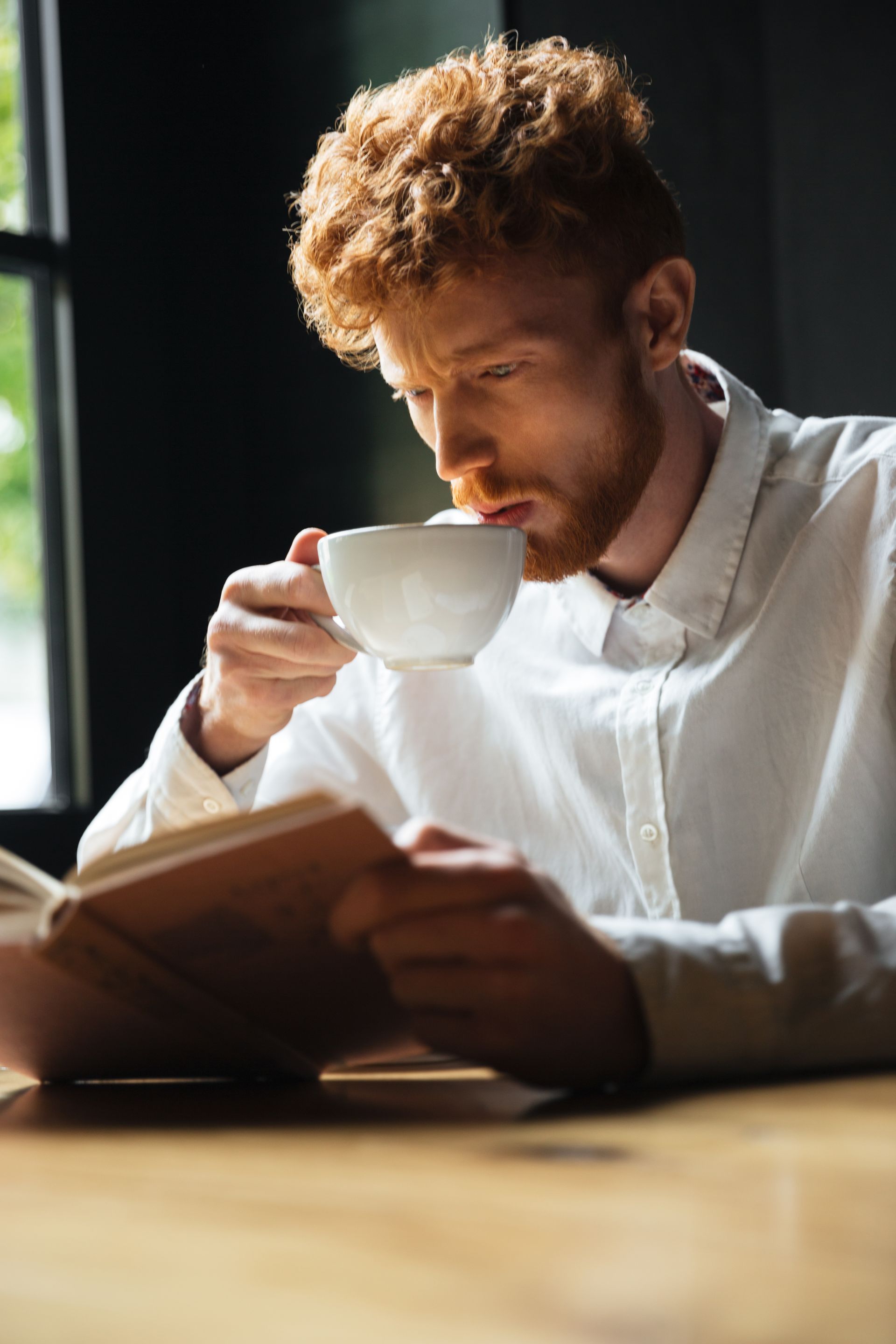 Person reading a book and sipping from a white cup by a sunlit window