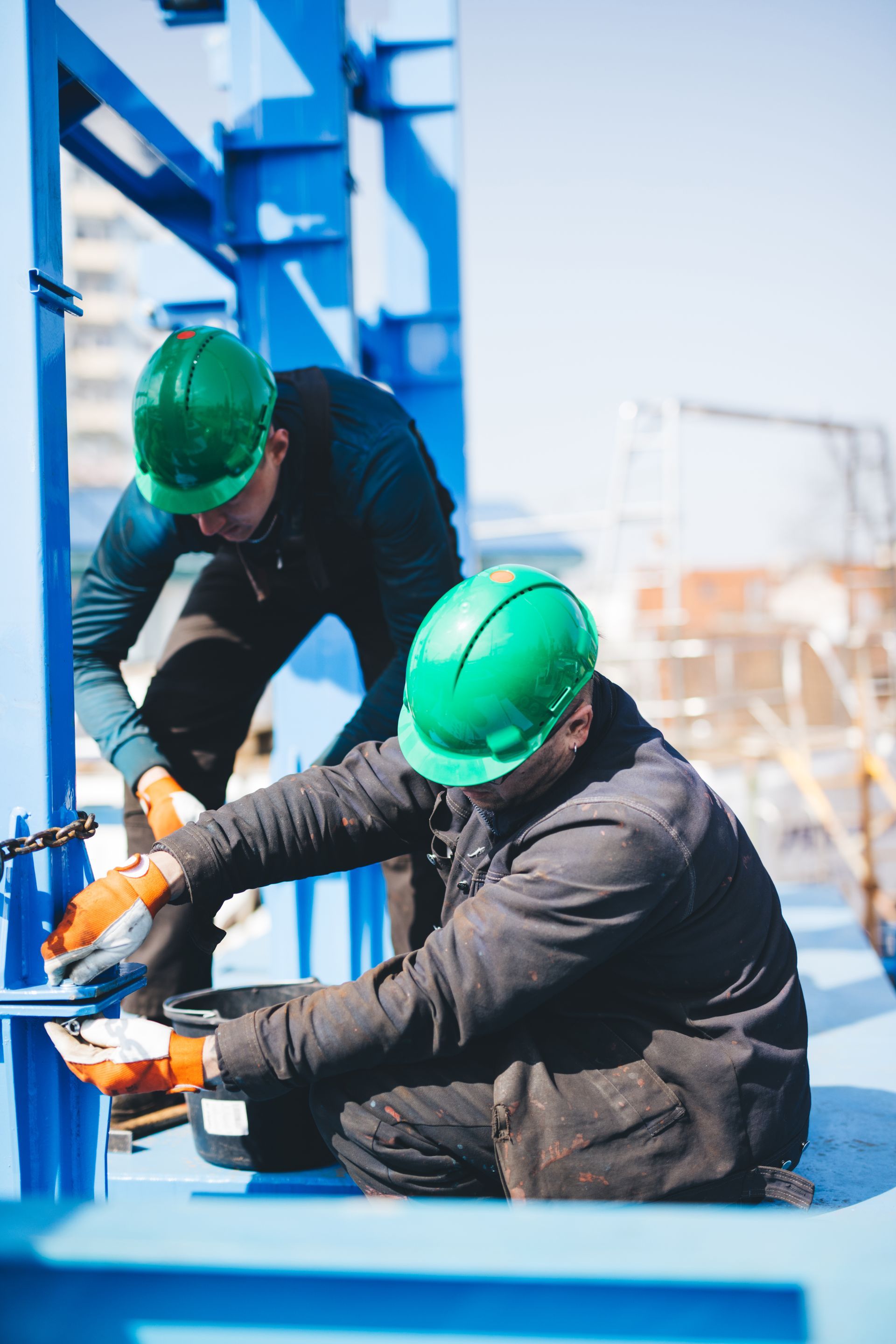 Two construction workers in green hard hats tightening bolts on a blue steel structure