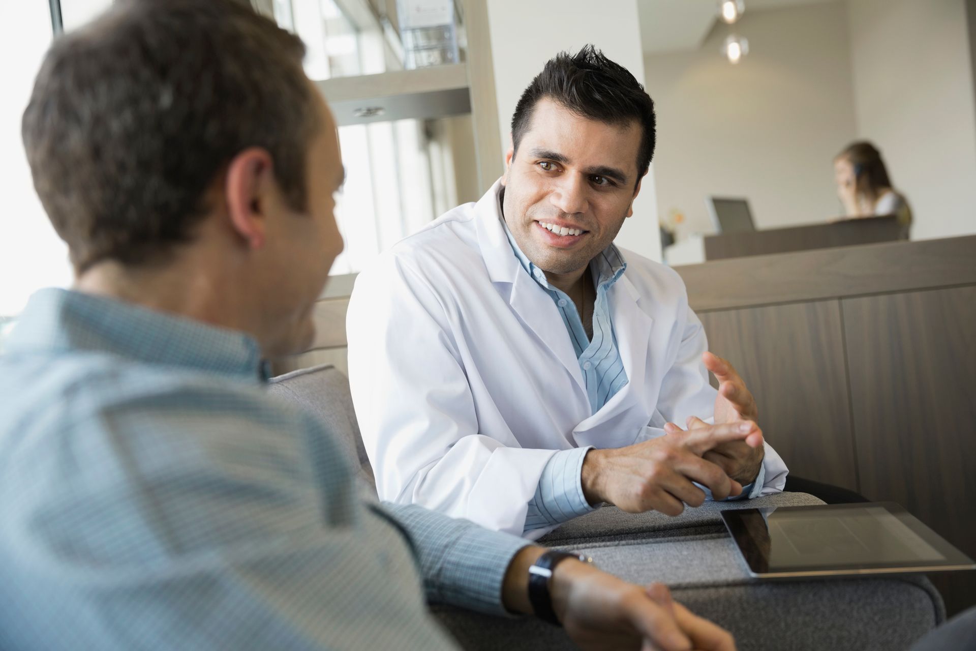 Two people talking in a modern office lounge, one in a white coat leaning forward and listening attentively.