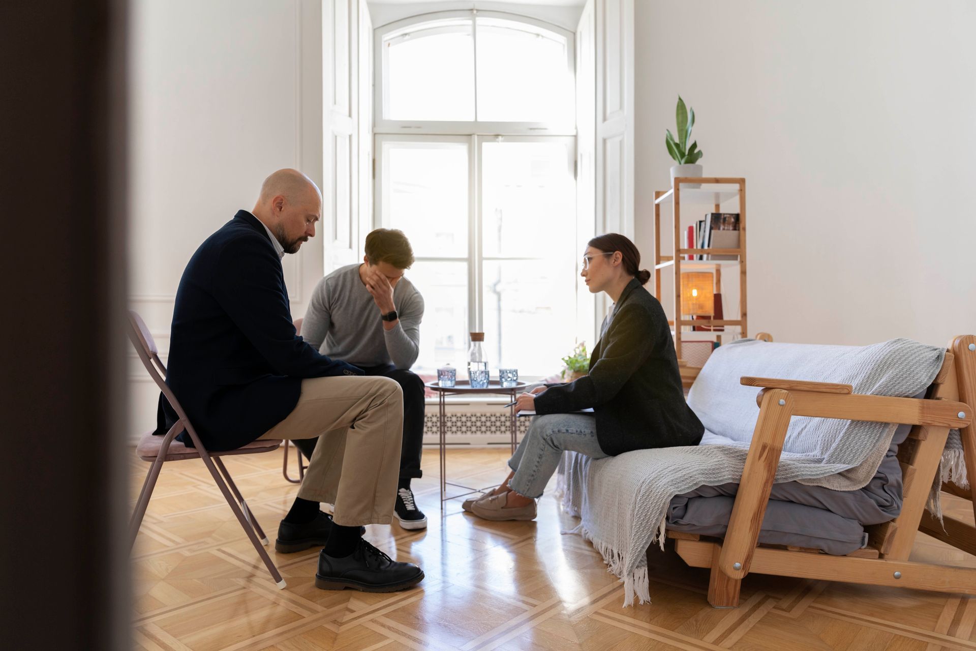 Three people talking in a bright living room, seated on chairs and a couch near a window.