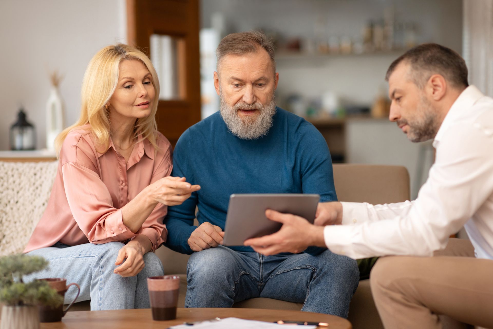 Three people discuss a tablet on a couch in a bright living room.