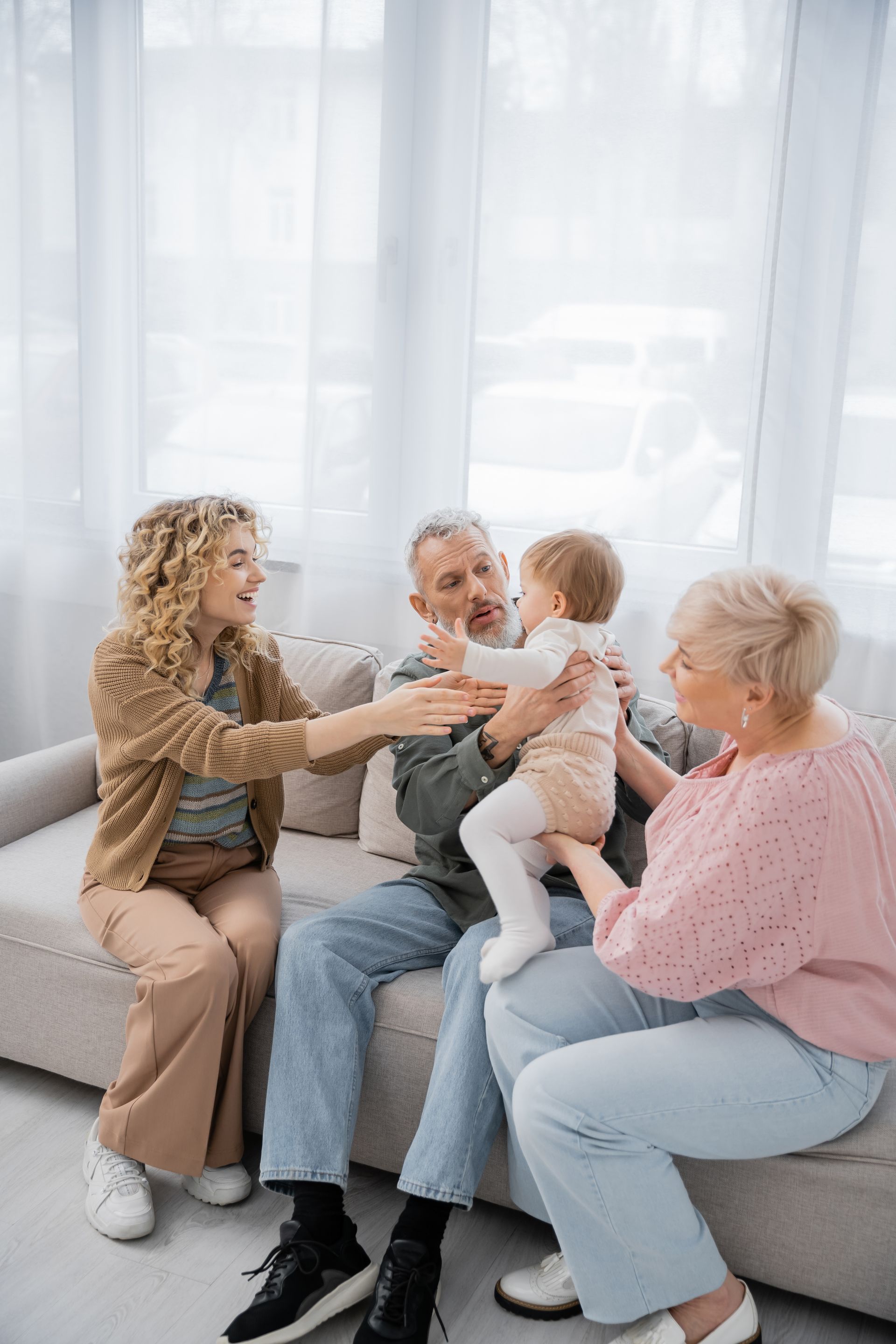 Four people sit on a couch, smiling and holding a baby in a bright living room.