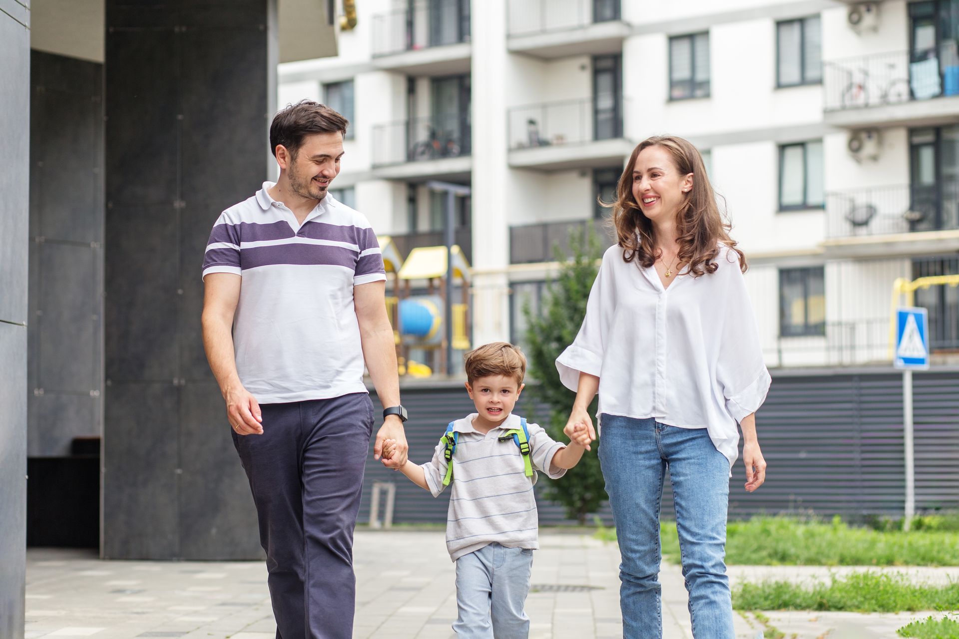 Three people walking on a city sidewalk, smiling and holding hands, with apartment buildings behind them.
