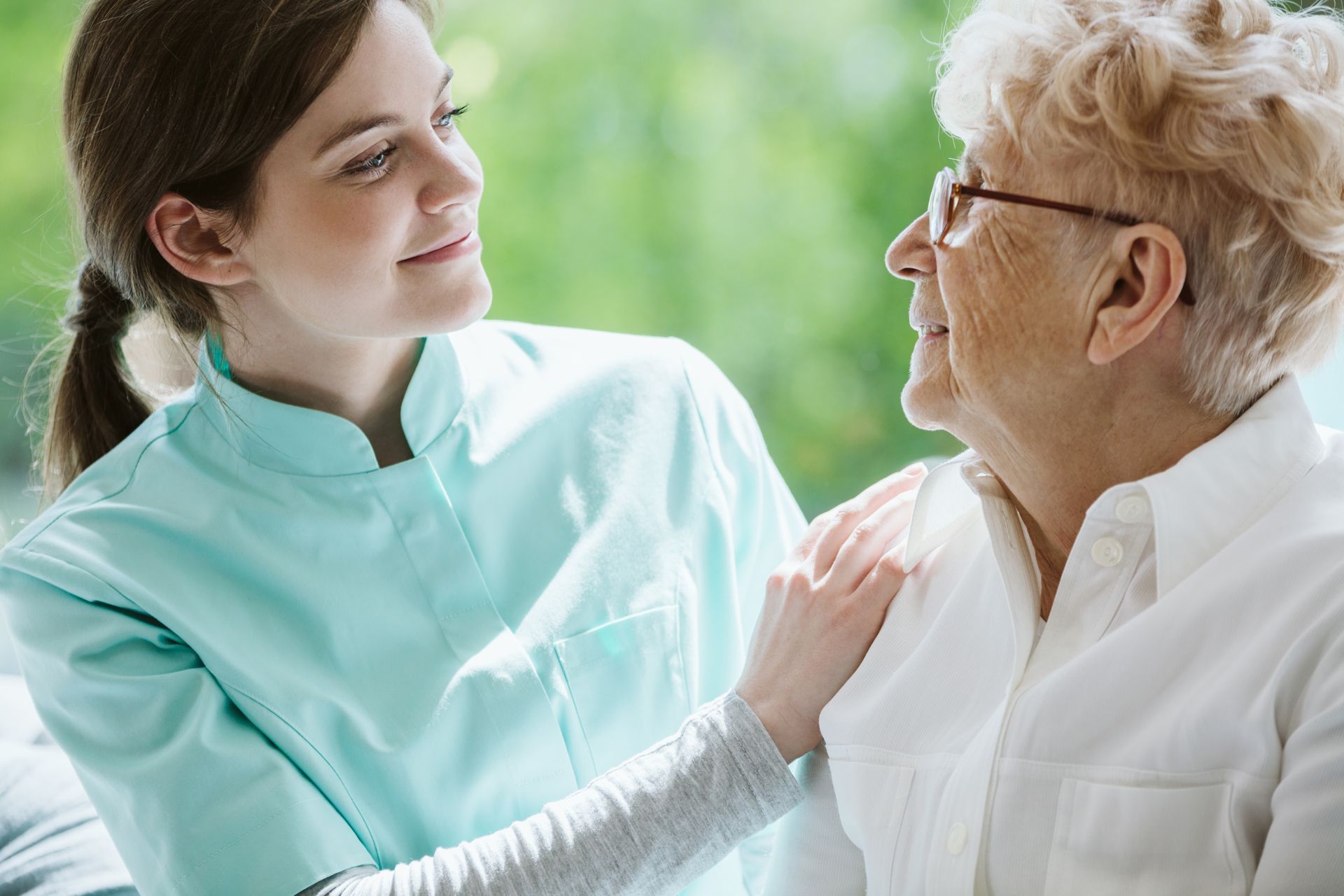 Caregiver in light blue uniform speaking with an elderly woman indoors near a bright window
