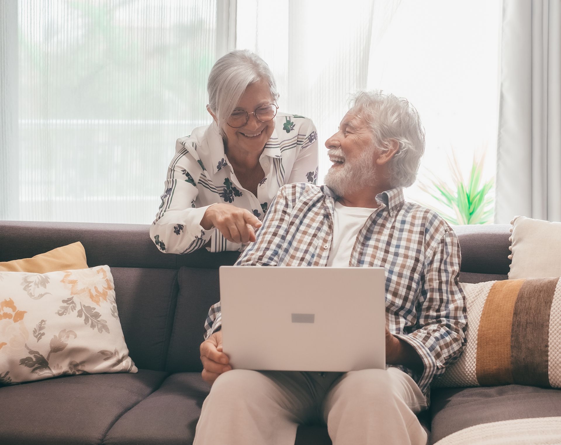 Two seniors smiling on a couch, sharing a laptop in a bright living room