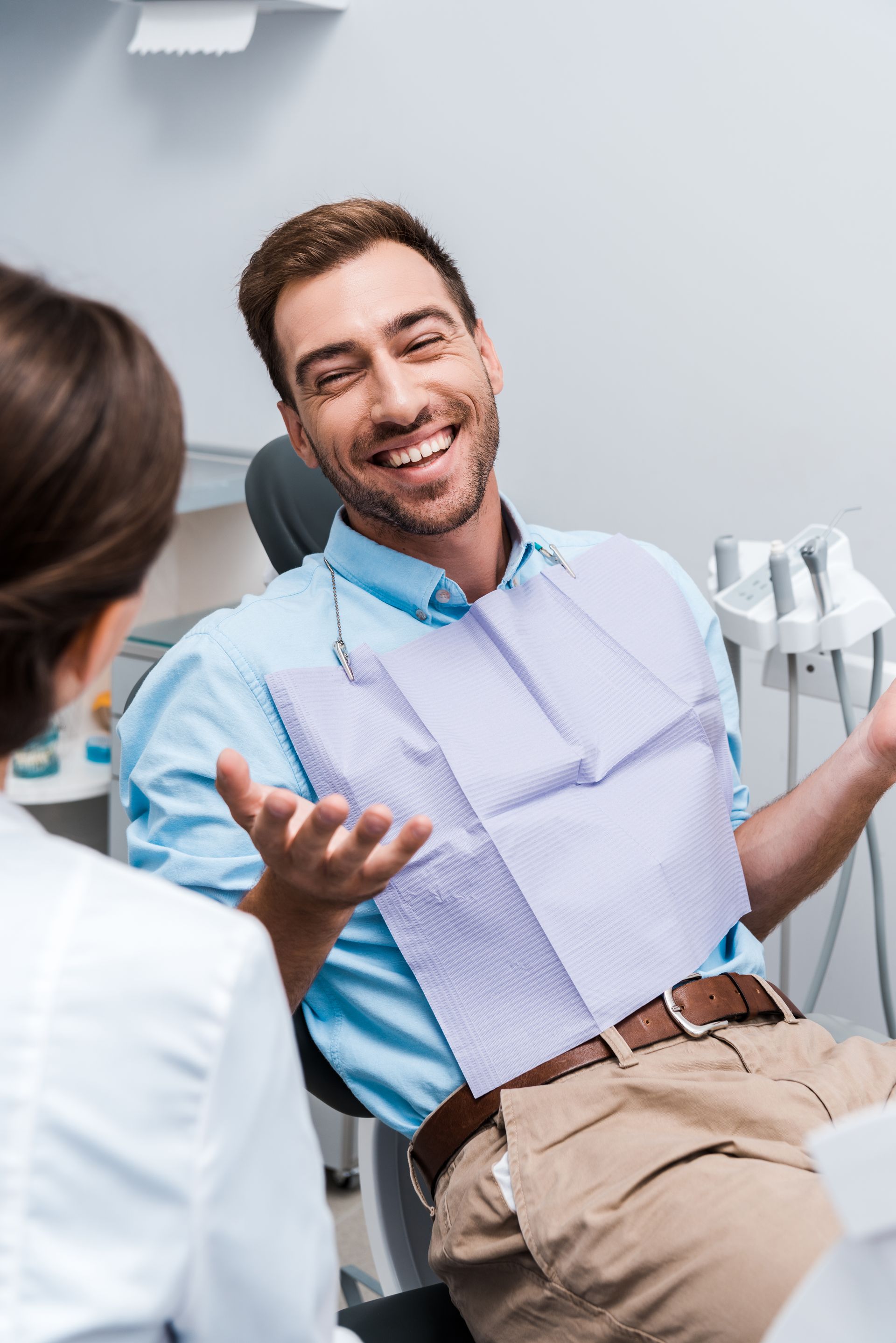Smiling man in blue shirt and bib in a dental chair, talking with a dentist in a clinic
