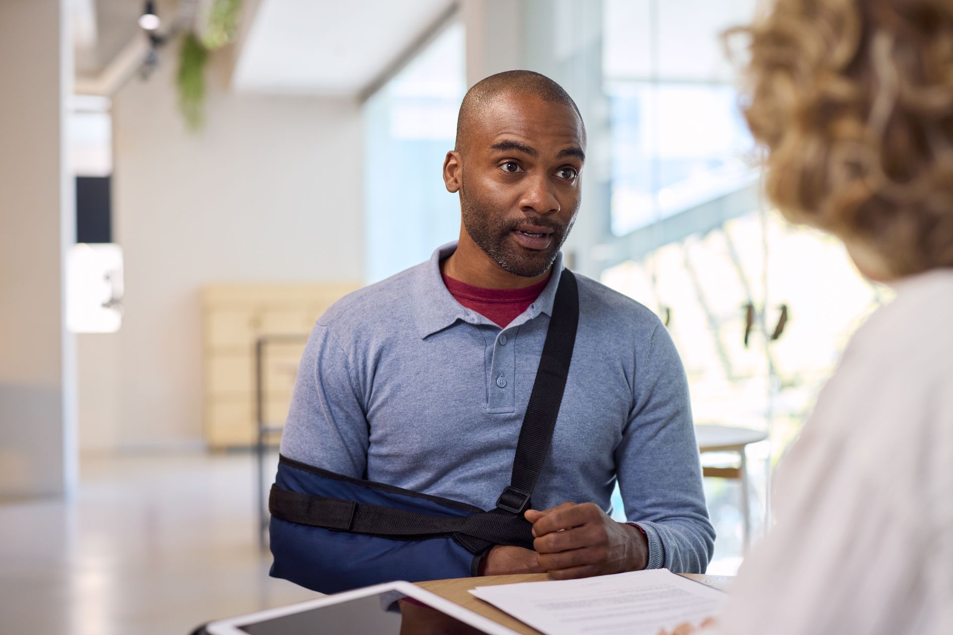Man in a blue sweater with arm in a sling speaking with someone at a desk in a bright office